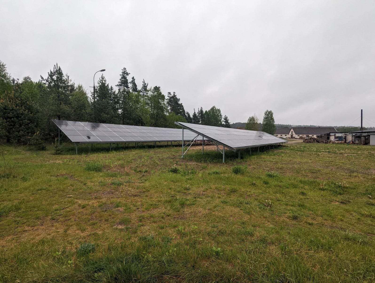 Dwie rzędy paneli fotowoltaicznych na metalowych konstrukcjach, na zewnątrz, na trawiastym terenie, w tle budynki gospodarcze i las, pochmurne niebo.