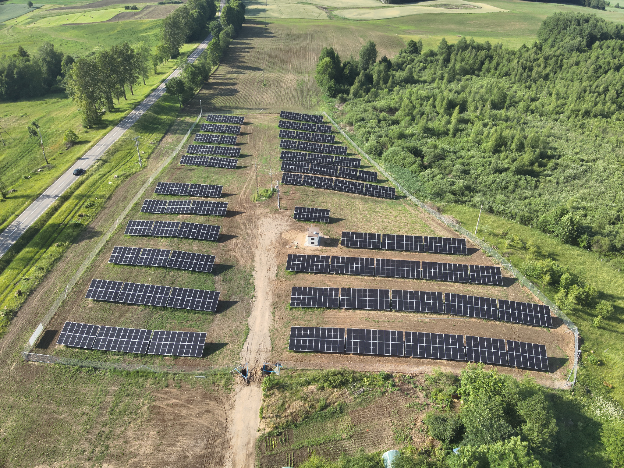 Widok z góry na farmę fotowoltaiczną z rzędami paneli na zielonym terenie, z drogą i lasem w tle. Widoczny mały budynek na terenie farmy i ogrodzenie.