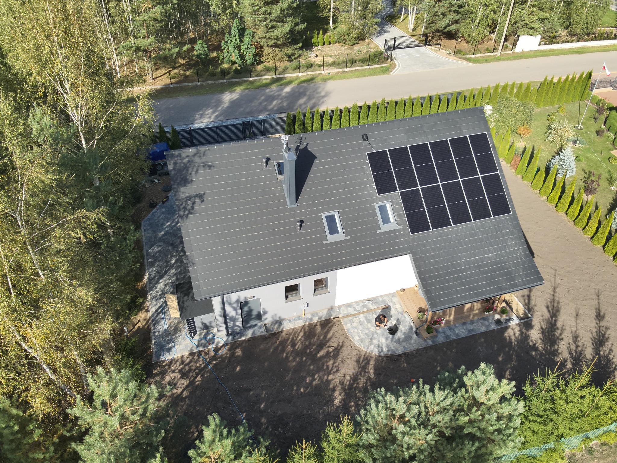 Widok z drona na dom z szarym dachem i panelami słonecznymi. Otoczenie to zadbany ogród z tujami i drzewami. Na podwórku widoczne świeżo ułożone kostki.