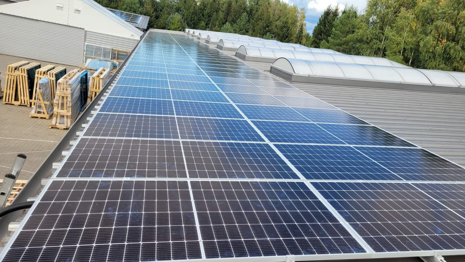 Panele fotowoltaiczne na dachu budynku przemysłowego, z paletami paneli w tle, gotowe do montażu. Widok z góry na instalację.