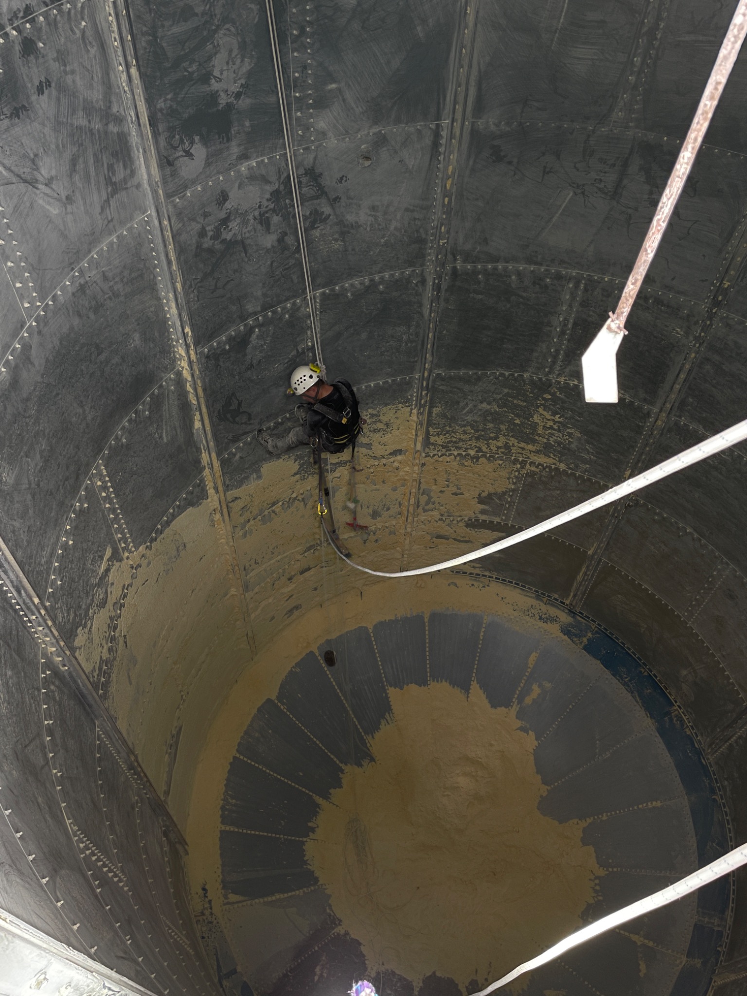 Pracownik na linach, w hełmie, czyści wnętrze zbiornika. Widok z góry ukazuje postęp prac, z wyraźną różnicą między czystą a zabrudzoną powierzchnią.