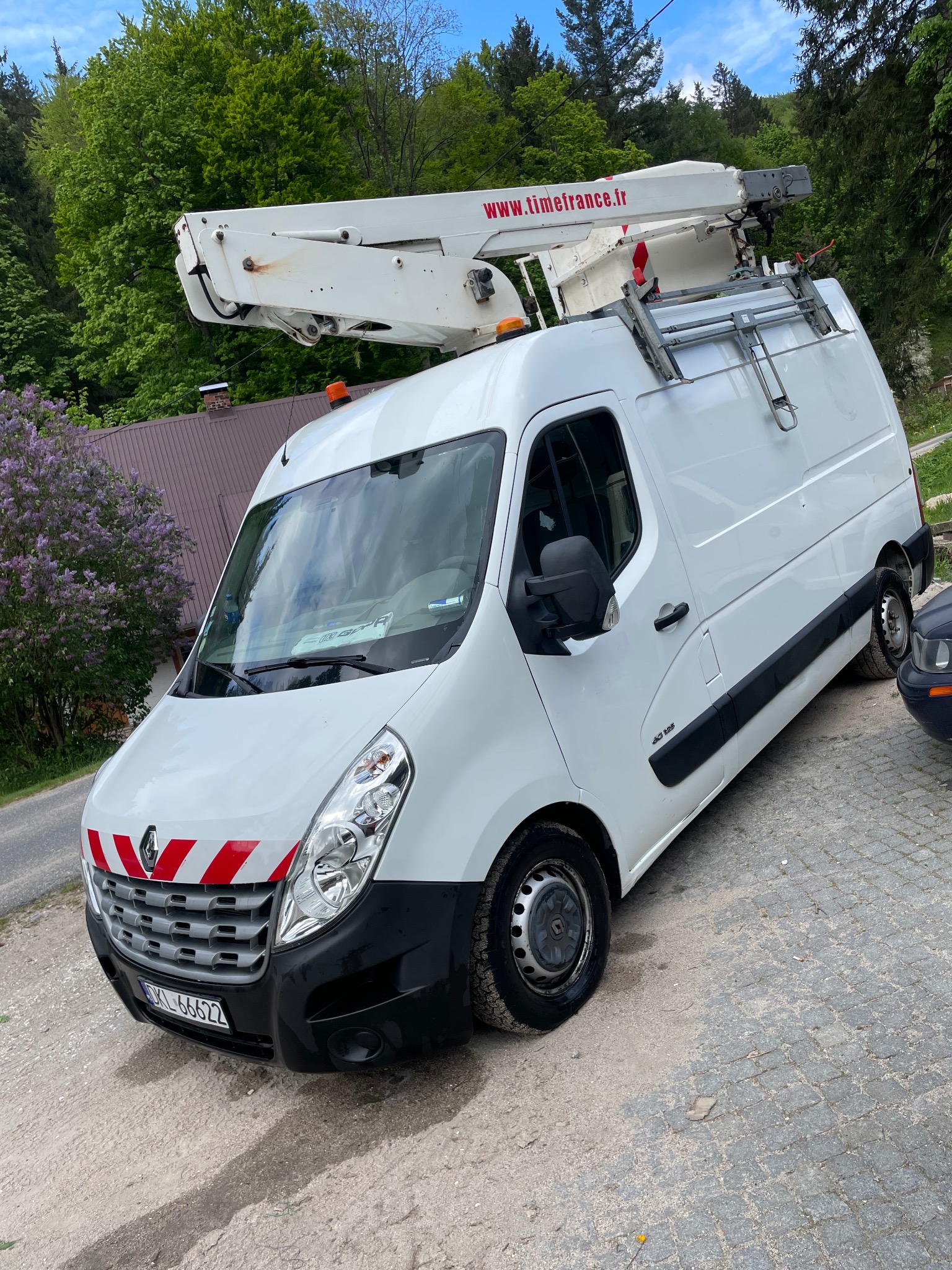 Biały furgon Renault z podnośnikiem koszowym Timefrance.fr na tle zieleni i błękitnego nieba. Pojazd zaparkowany na kostce brukowej, widoczne tablice rejestracyjne.