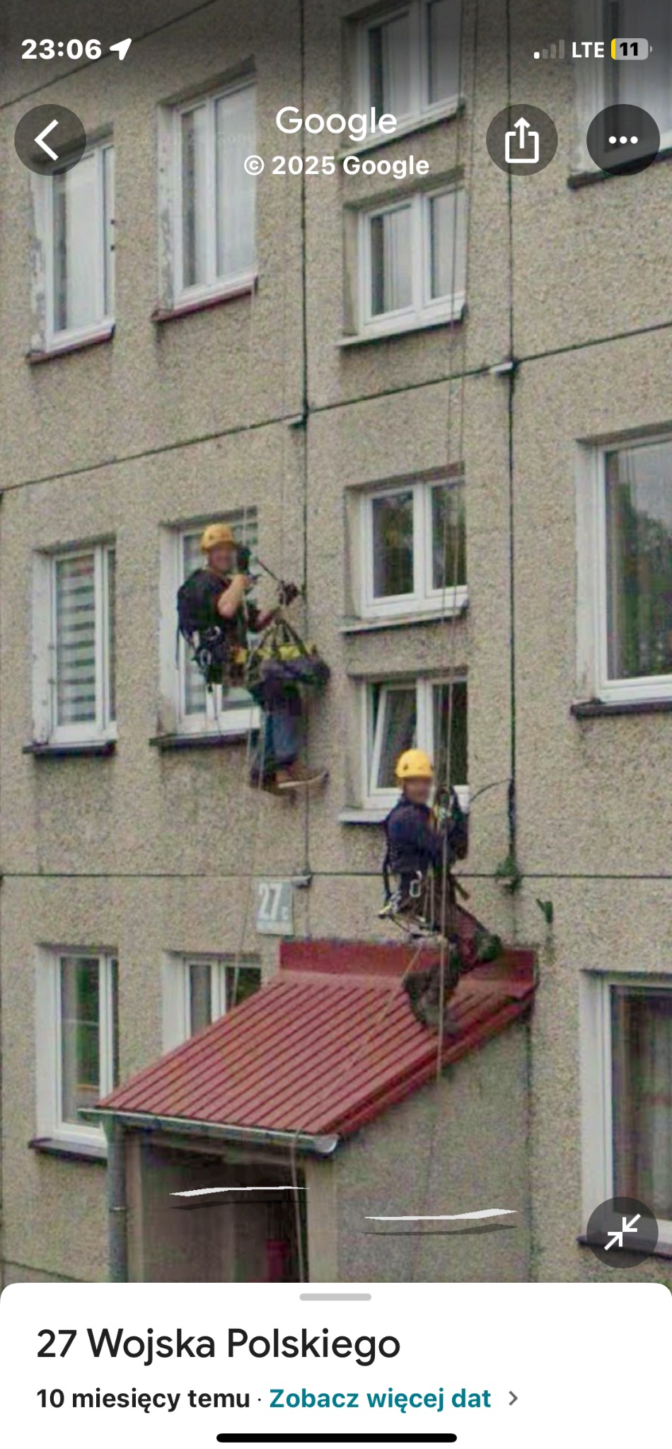 Dwóch pracowników wysokościowych w kaskach, na linach, przy elewacji bloku. Jeden z narzędziami, drugi nad wejściem z czerwonym zadaszeniem. Widok z dołu.