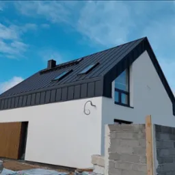 Nowoczesny dom z ciemnym, metalowym dachem i oknami dachowymi, na tle błękitnego nieba z chmurami. Widoczne detale wykończenia i elementy konstrukcyjne.