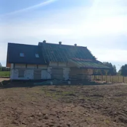 Dom w trakcie budowy z częściowo położonym ciemnym dachem i widoczną więźbą. Na pierwszym planie ziemia, w tle błękitne niebo z jasnym słońcem.