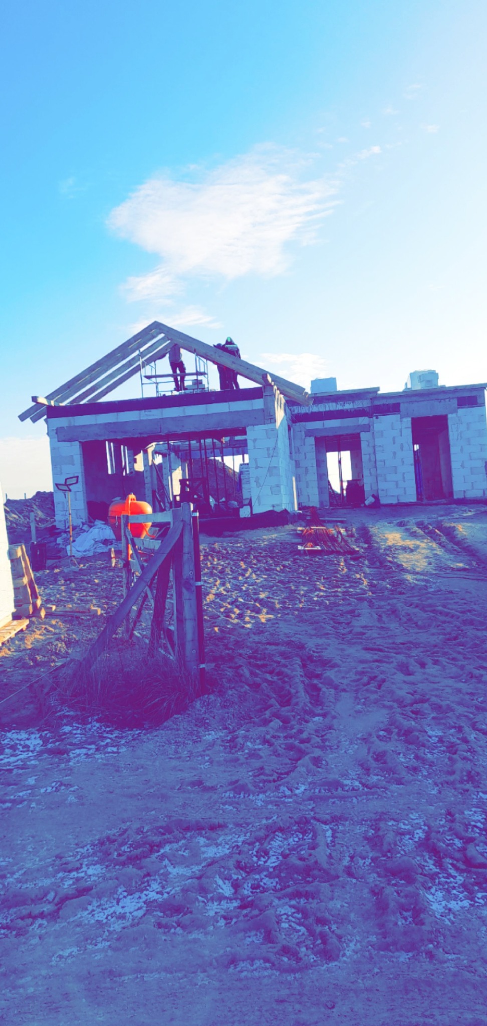 Wznoszenie więźby dachowej na surowym budynku z białego bloczka, widok z dołu na tle błękitnego nieba. Na dachu robotnicy w trakcie prac.