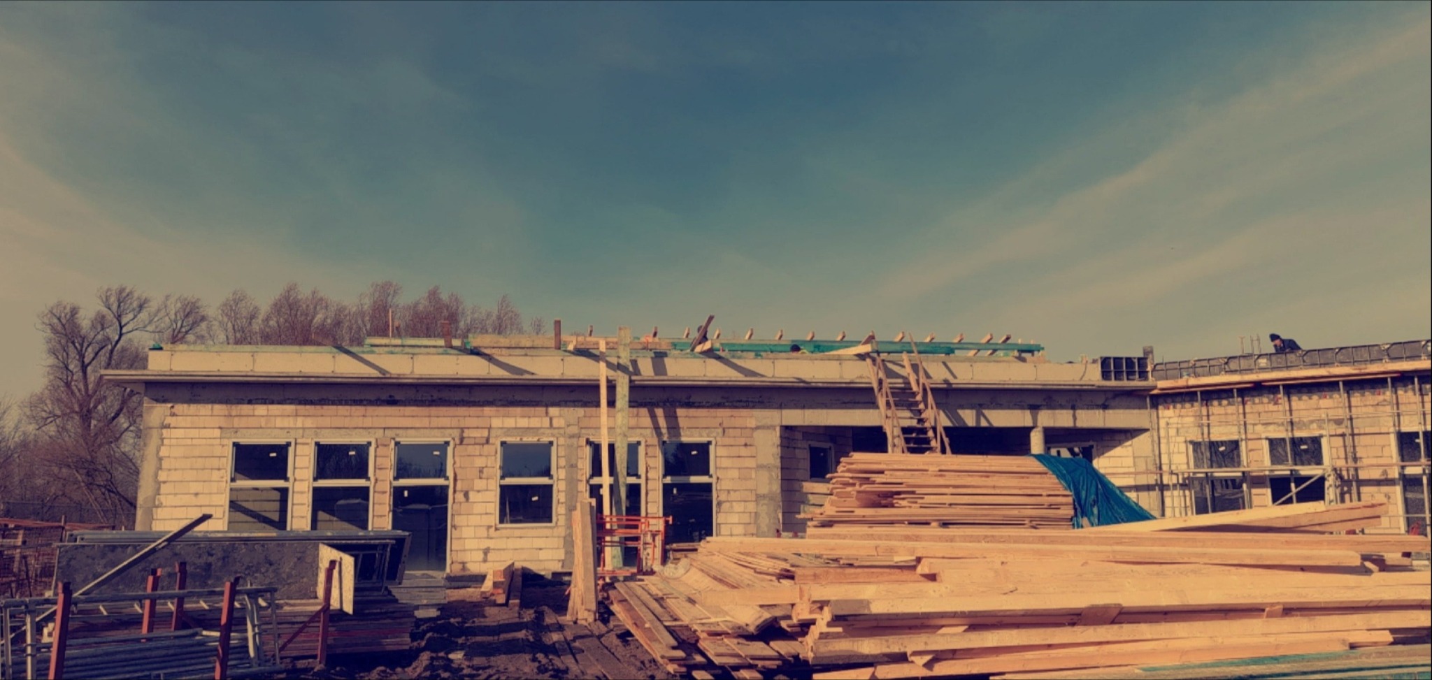 Budynek w trakcie budowy dachu, widoczne drewniane elementy konstrukcyjne i sterta desek na pierwszym planie. Na dachu widoczna drabina i osoba.