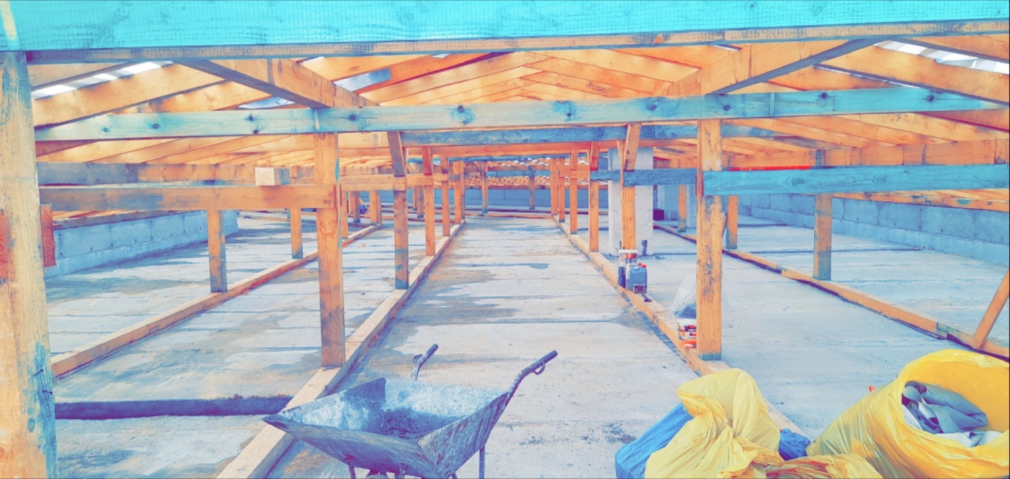 Wnętrze budowy z drewnianą konstrukcją dachu, widoczne pustaki i wózek budowlany. Elementy więźby dachowej w surowym stanie przed dalszymi pracami.