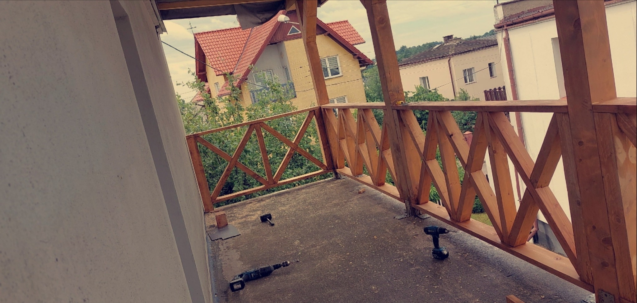 Drewniana balustrada balkonu w trakcie montażu, widoczne narzędzia: wiertarka, młotek. W tle zabudowa jednorodzinna i zieleń.