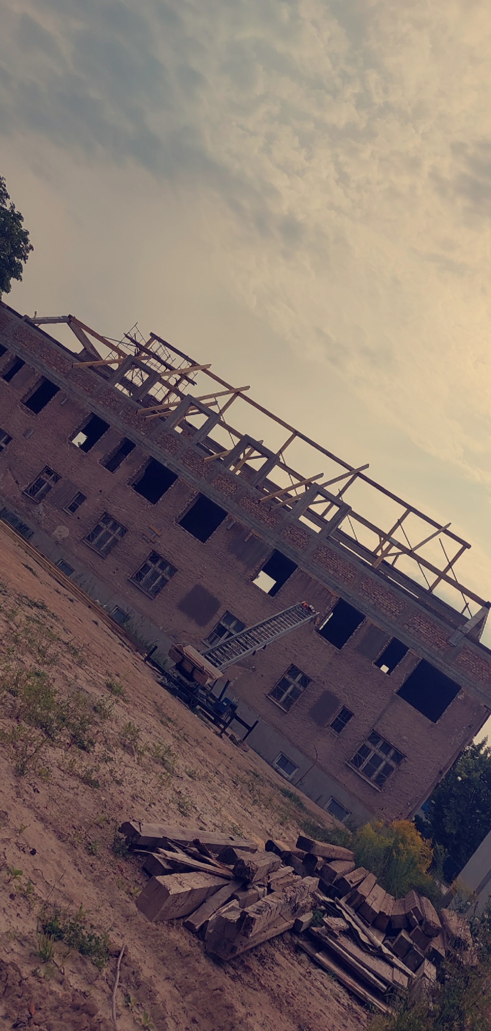 Budynek w trakcie budowy dachu, widoczna więźba i ceglana konstrukcja. Na ziemi leżą materiały budowlane. Ustawiona drabina.