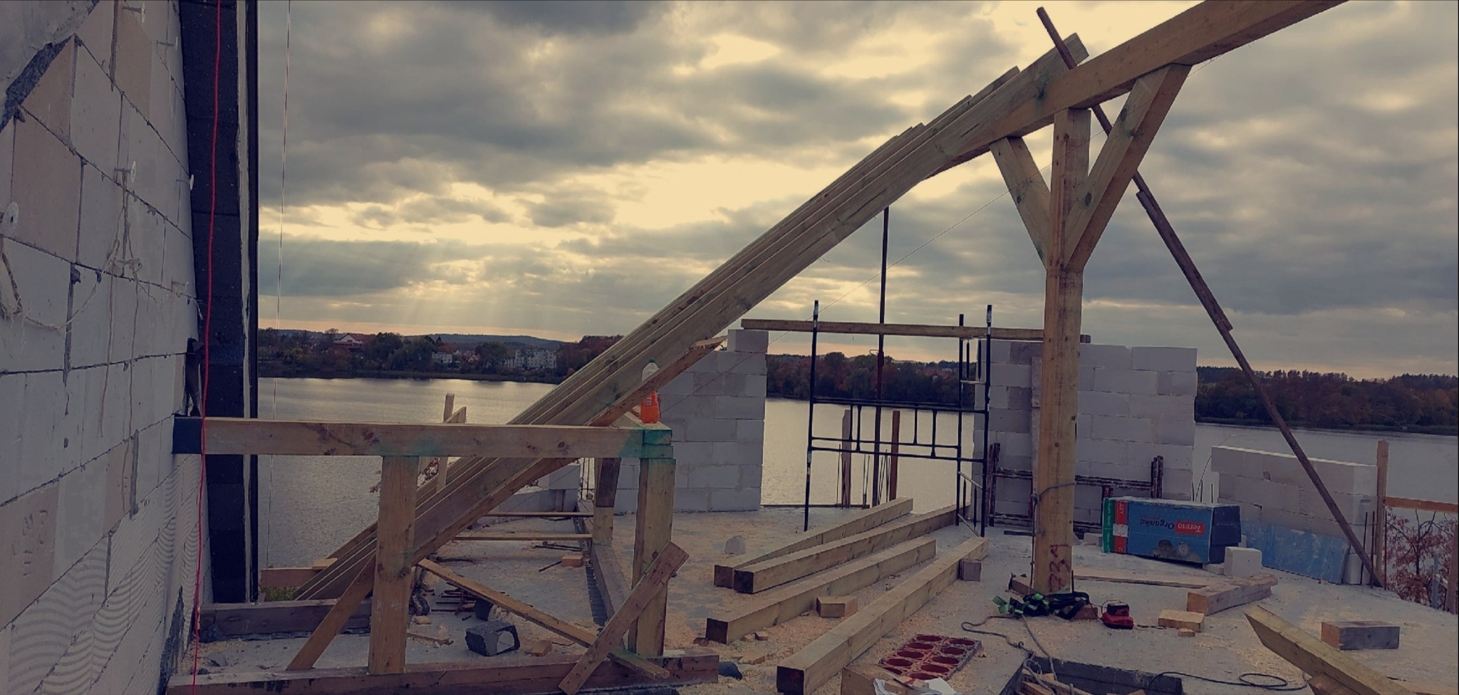 Wznoszenie drewnianej konstrukcji dachu na budynku z bloczków, z malowniczym widokiem na jezioro w tle, przy pochmurnym niebie. Elementy więźby dachowej na placu budowy.