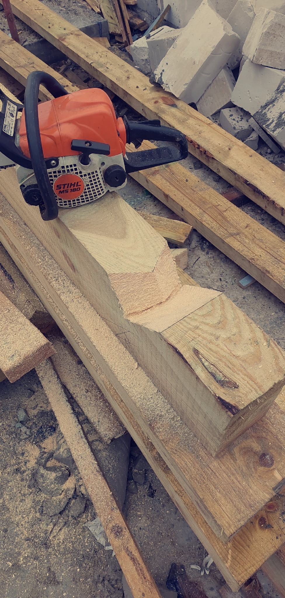 Pomarańczowa piła łańcuchowa Stihl MS 180 na drewnianej belce z wyciętym fragmentem, w otoczeniu drewnianych desek i gruzu. Przygotowanie elementu więźby dachowej.