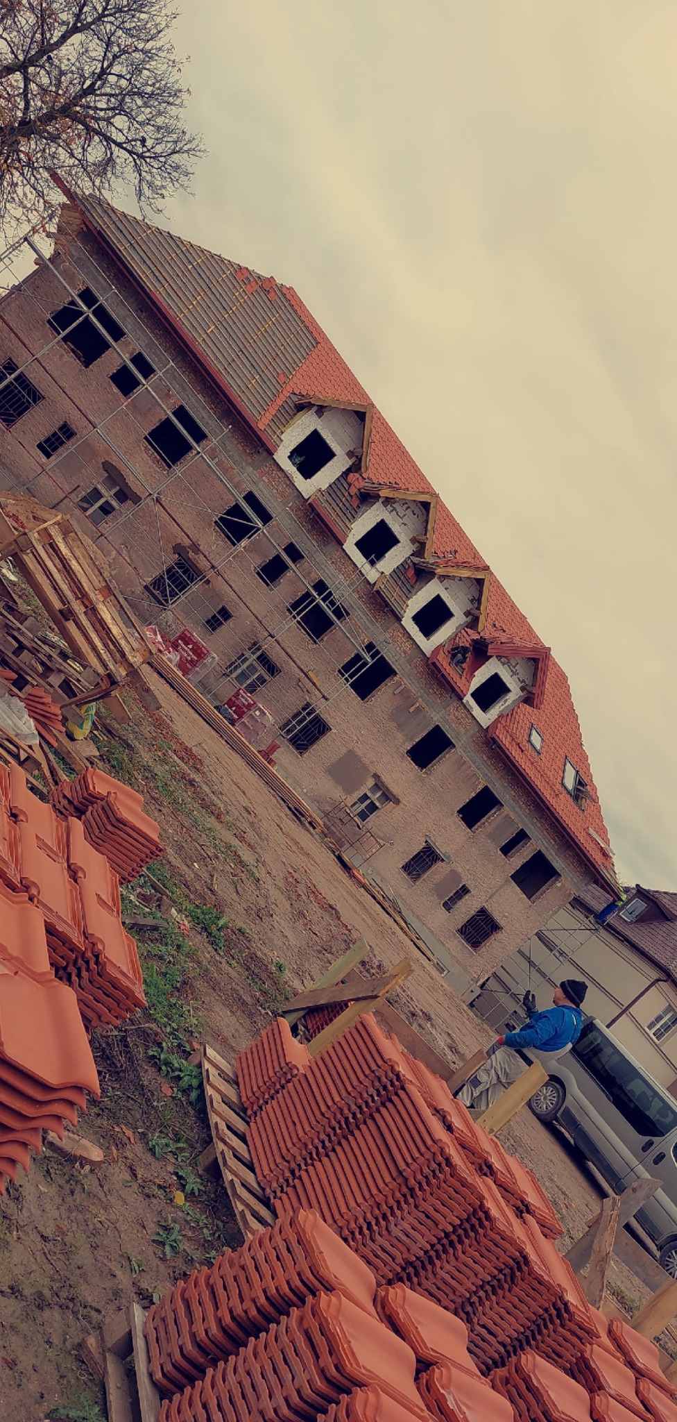 Ceglasty budynek w trakcie krycia dachówką, widoczne rusztowania i stosy dachówek. Pracownik w niebieskiej kurtce przy samochodzie. Ujęcie z dołu.