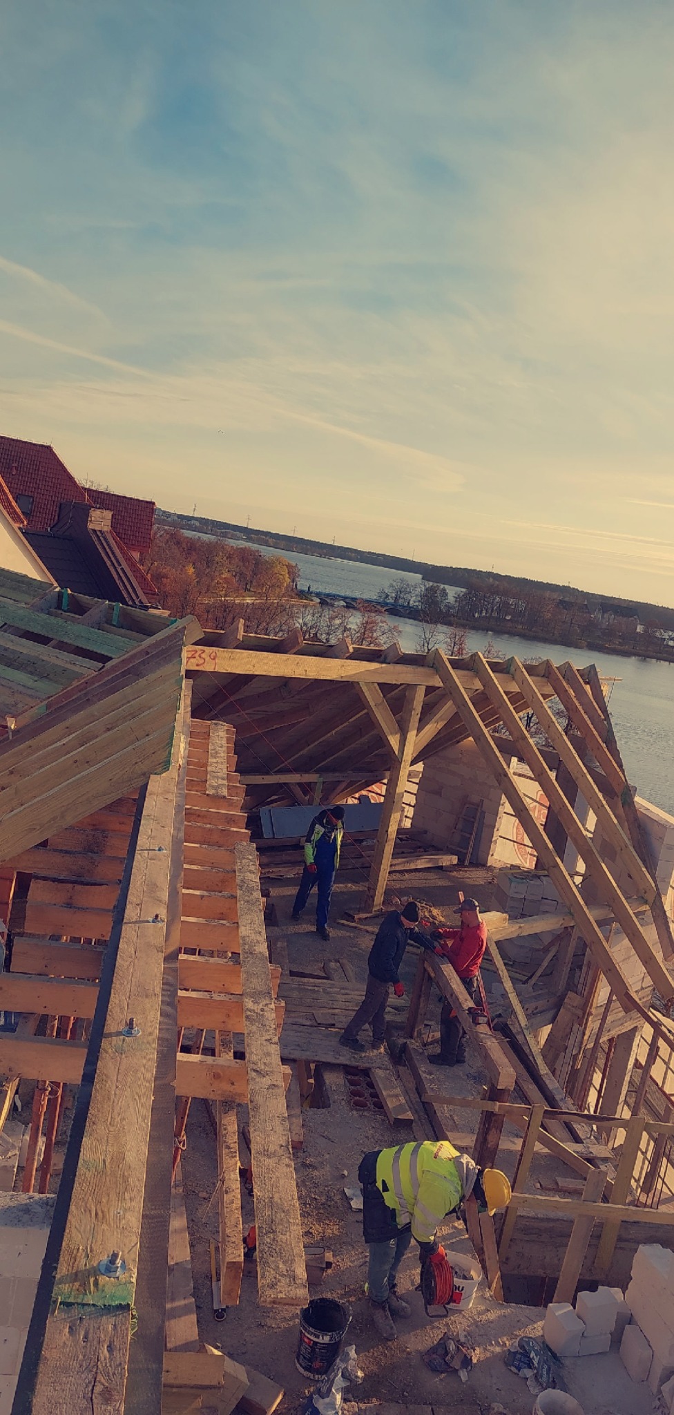 Wysoki kąt: ekipa montuje więźbę dachową na budynku z widokiem na rzekę. Konstrukcja z drewnianych belek i desek, robotnicy w odzieży roboczej. Częściowo pokryty dach.
