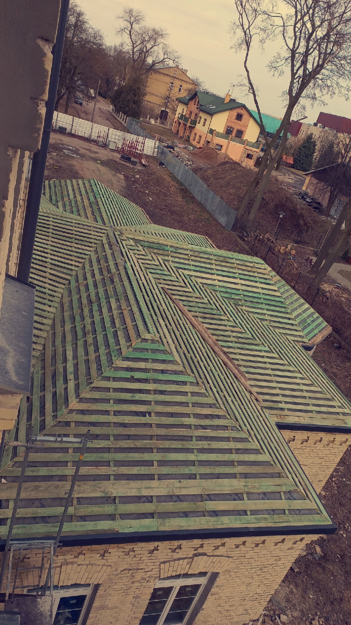 Konstrukcja dachu z łatami i kontrłatami, widok z góry na budynek z cegły, przygotowana pod pokrycie dachowe, w tle zabudowa jednorodzinna i drzewa.