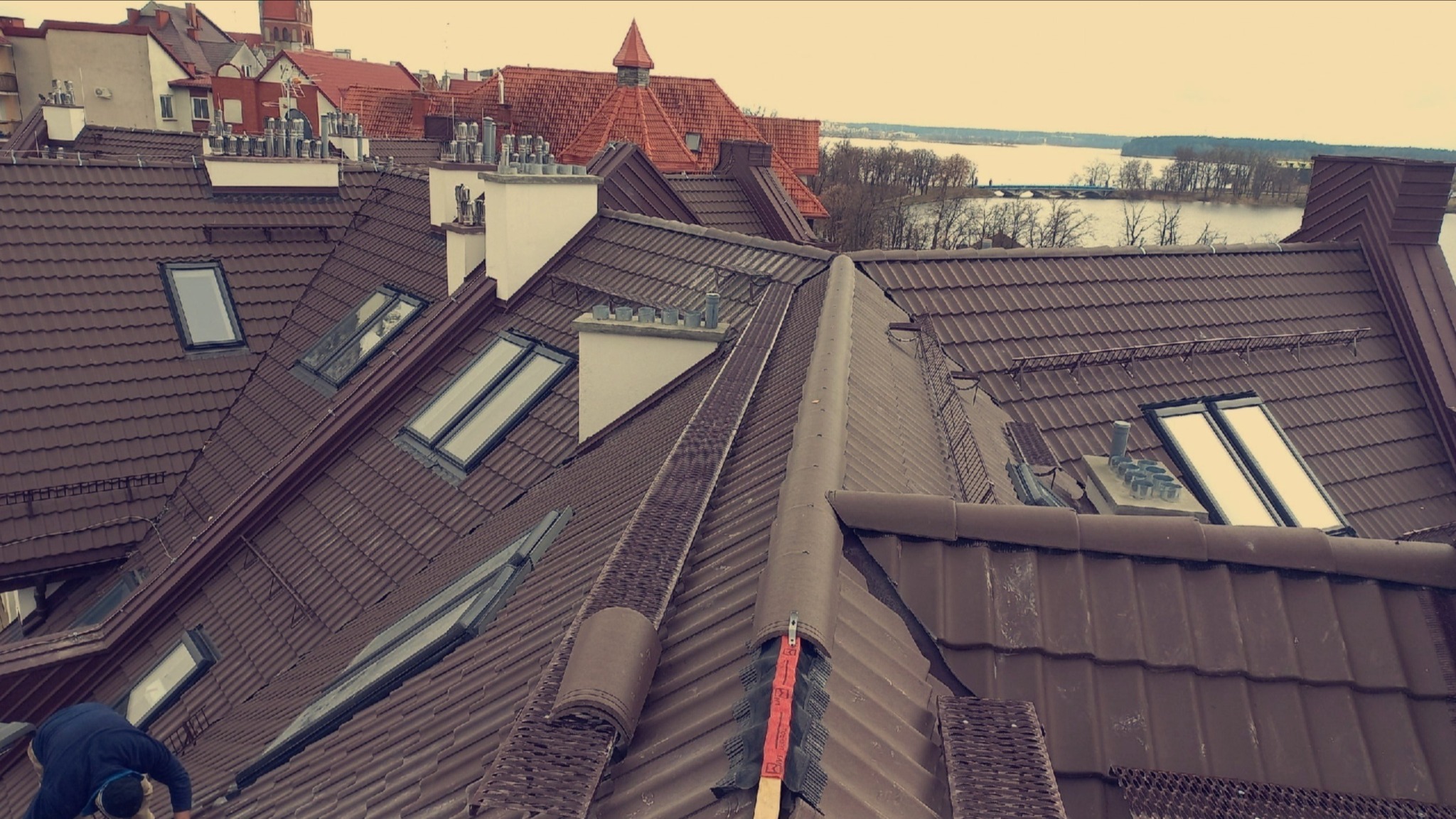Brązowy dach z dachówką ceramiczną i oknami połaciowymi na tle zabudowy miejskiej i jeziora. Widoczny dekarz podczas pracy.