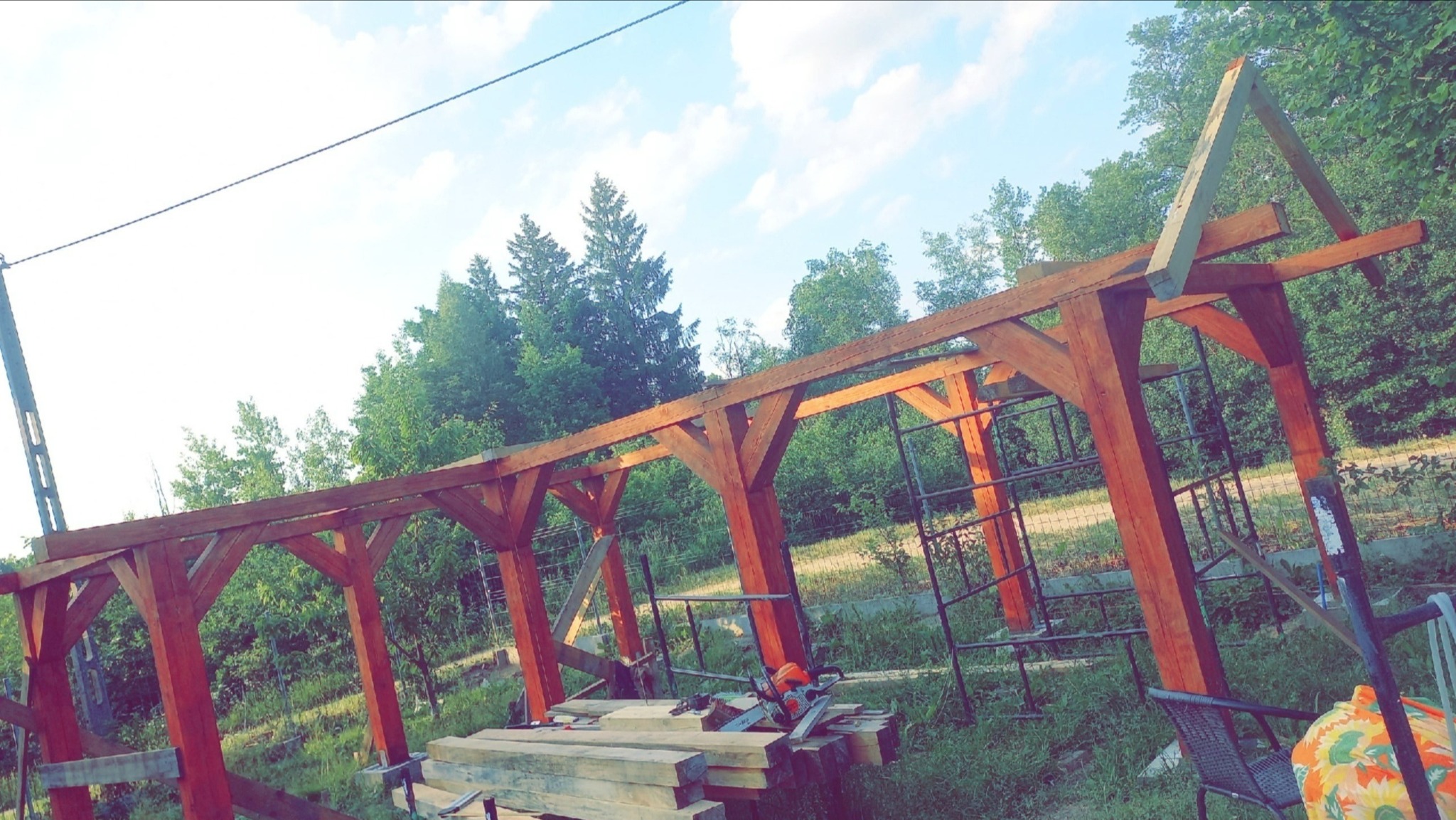Wznoszenie drewnianej konstrukcji z belek na tle zieleni, widoczne narzędzia. Etap budowy z elementami więźby dachowej w słoneczny dzień.