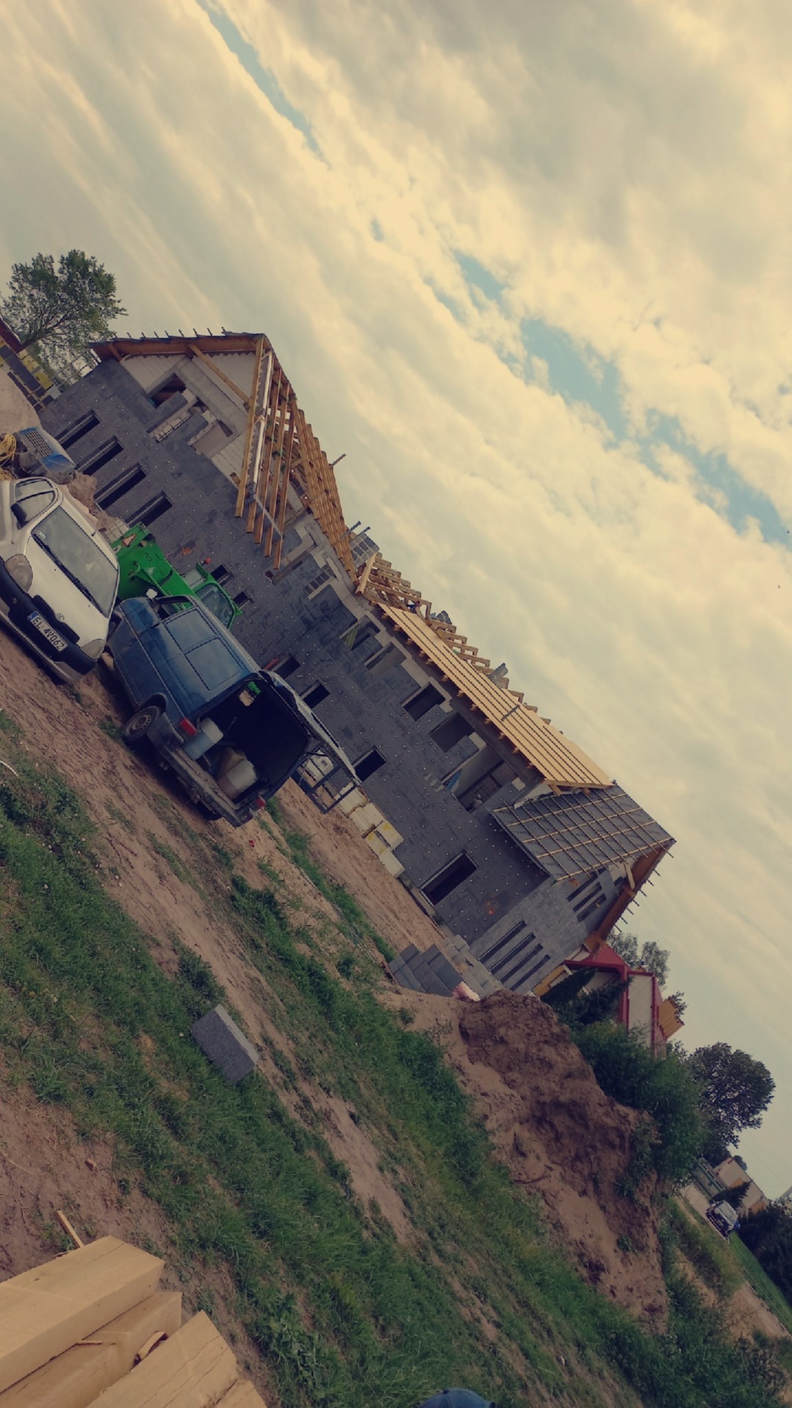 Wznoszony budynek wielorodzinny z częściowo wykonanym dachem i widoczną więźbą. Na pierwszym planie zaparkowane samochody i materiały budowlane.