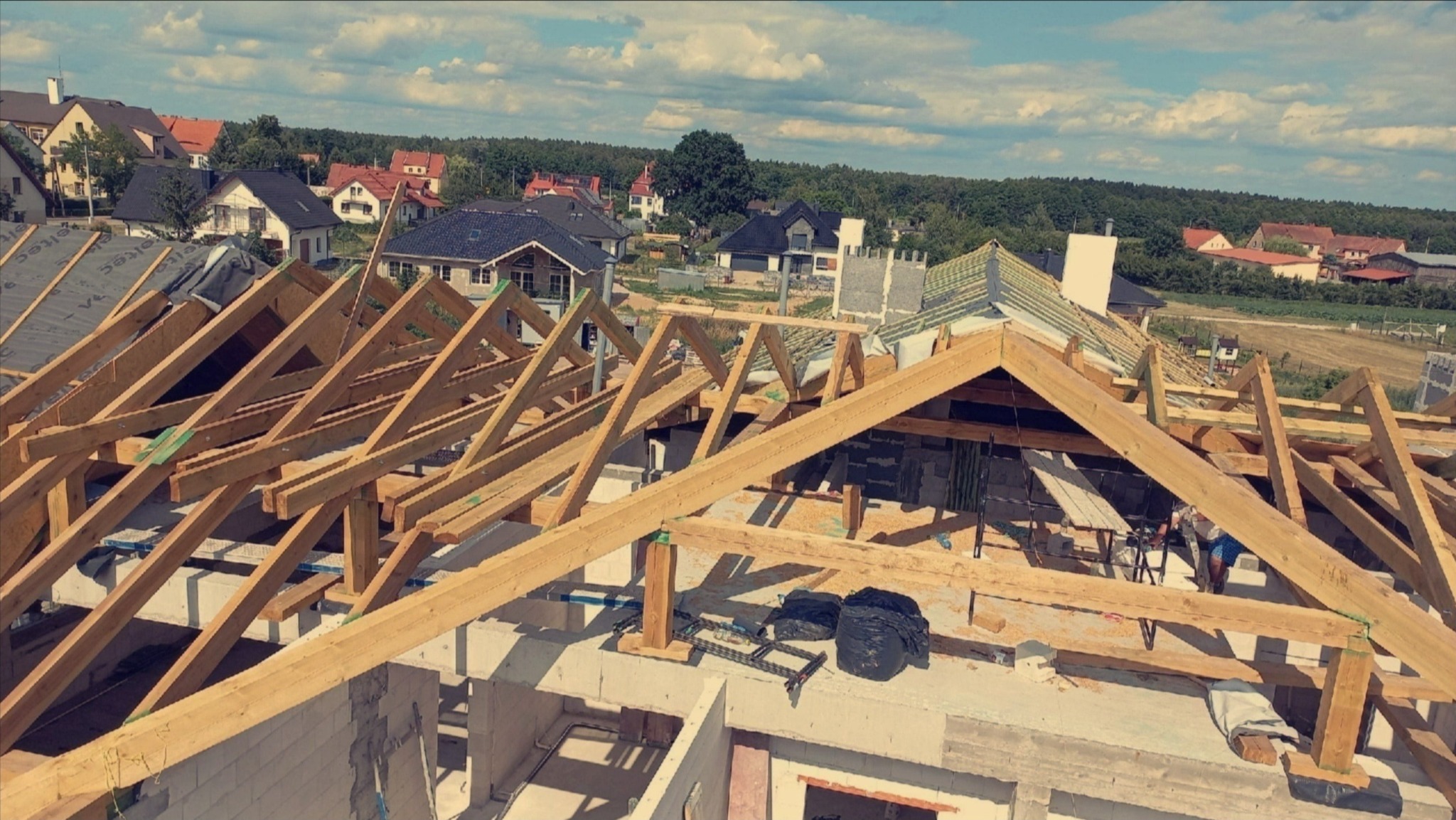 Wznoszenie drewnianej więźby dachowej na nowym budynku, widok z góry na konstrukcję w trakcie montażu, w tle zabudowa jednorodzinna i zieleń.