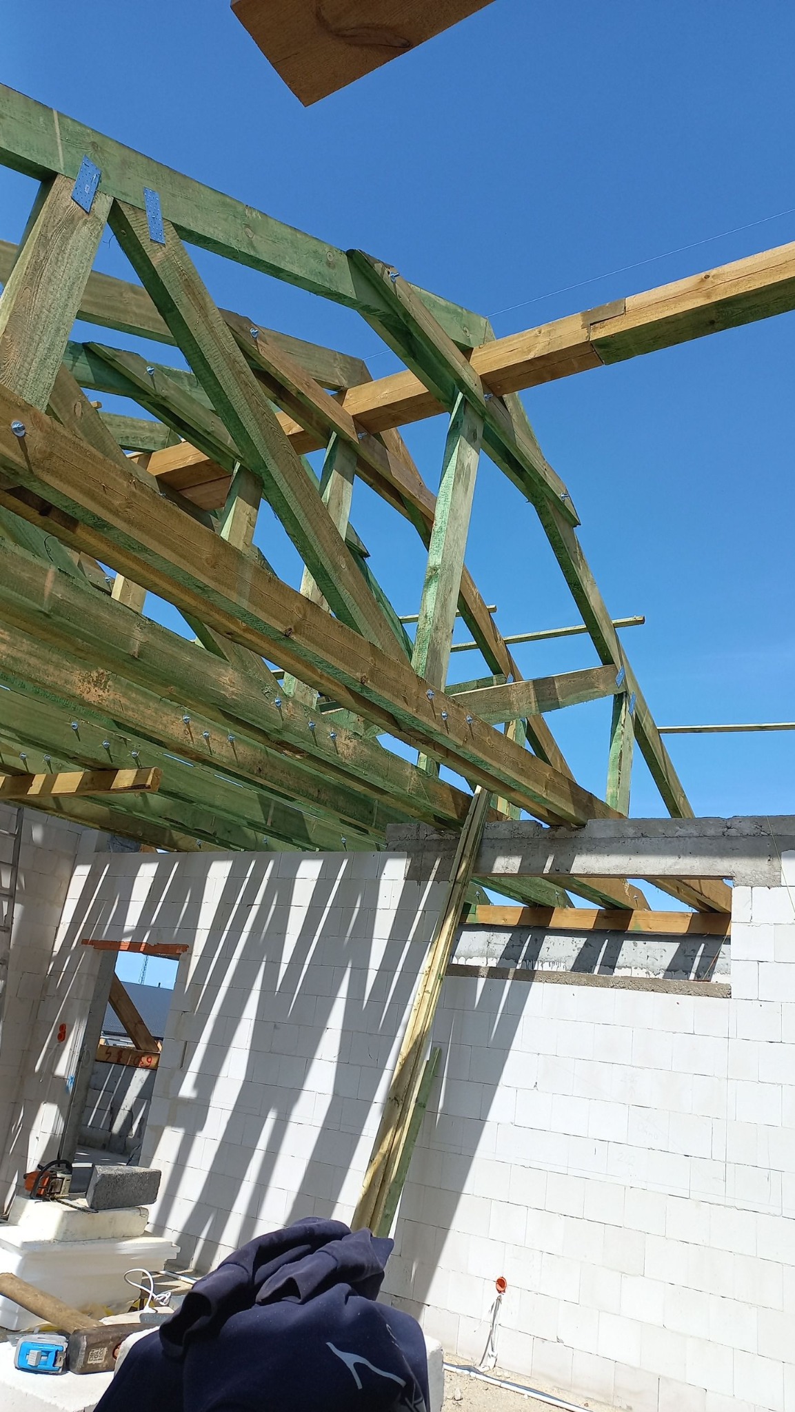 Drewniana konstrukcja więźby dachowej z zielonkawą impregnacją na tle błękitnego nieba, widoczna z wnętrza budynku w trakcie budowy, z elementami z betonu komórkowego.