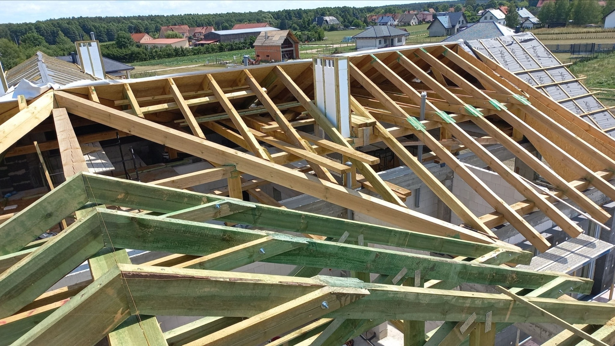 Wznoszenie drewnianej więźby dachowej z zielonymi elementami impregnacji, widok z góry na tle zabudowy jednorodzinnej i zieleni. Etap budowy dachu.