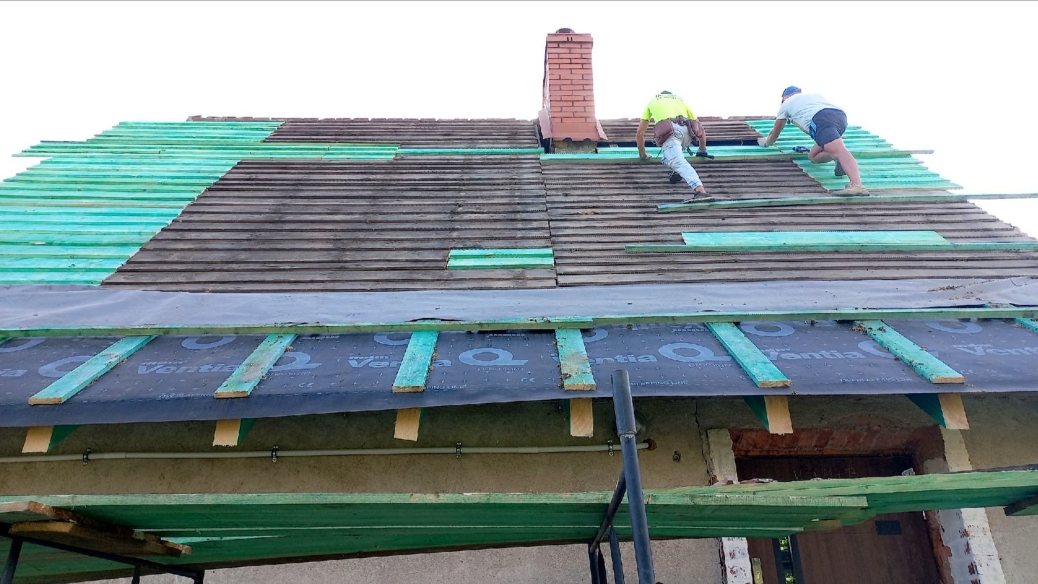 Dwóch dekarzy pracuje na skośnym dachu domu, układając nową warstwę izolacyjną i łaty pod pokrycie, widoczny komin. Częściowo zdjęte stare dachówki.