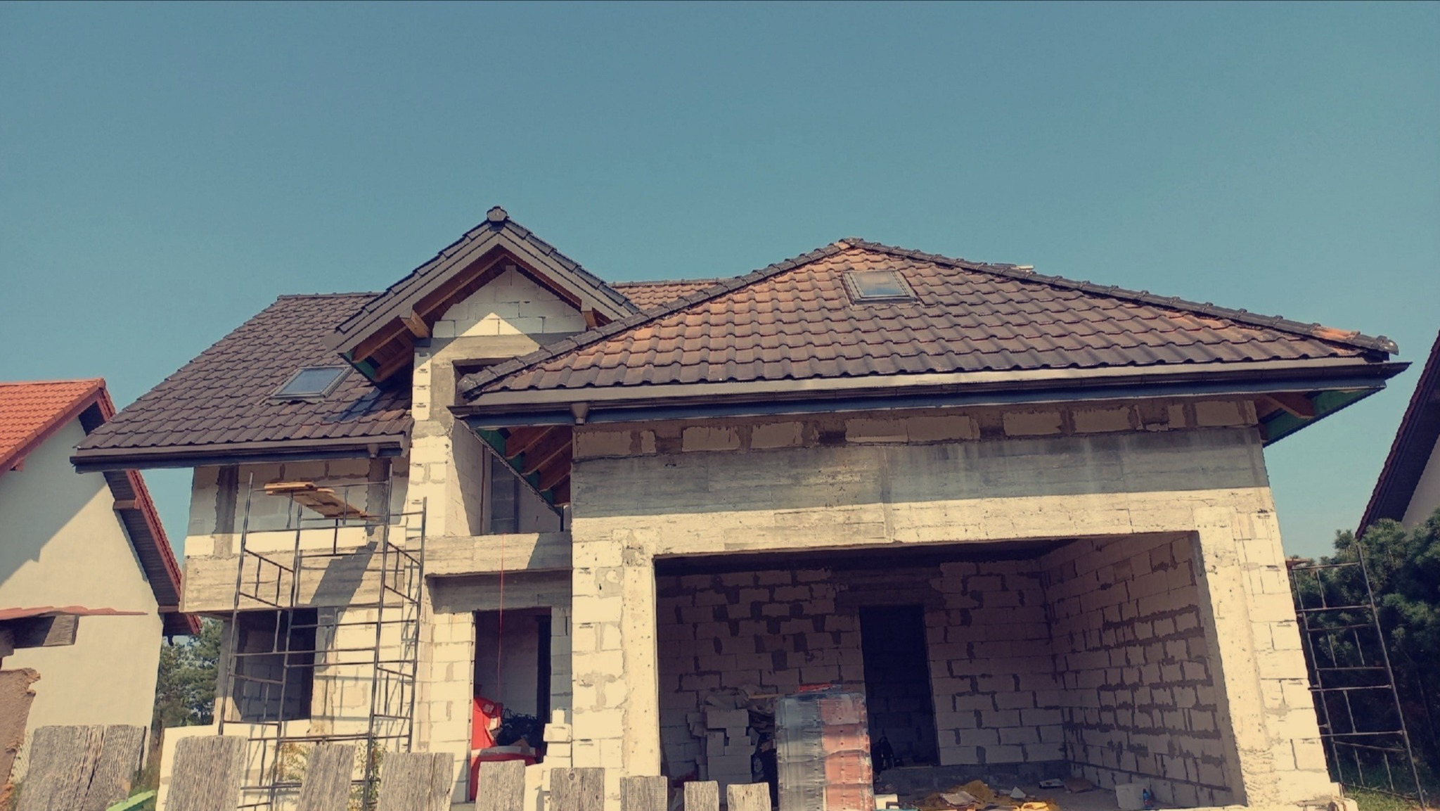 Dom w trakcie budowy z ciemną dachówką i zamontowanymi rynnami, widoczne rusztowania i elementy konstrukcyjne. Ujęcie z dołu, błękitne niebo w tle.