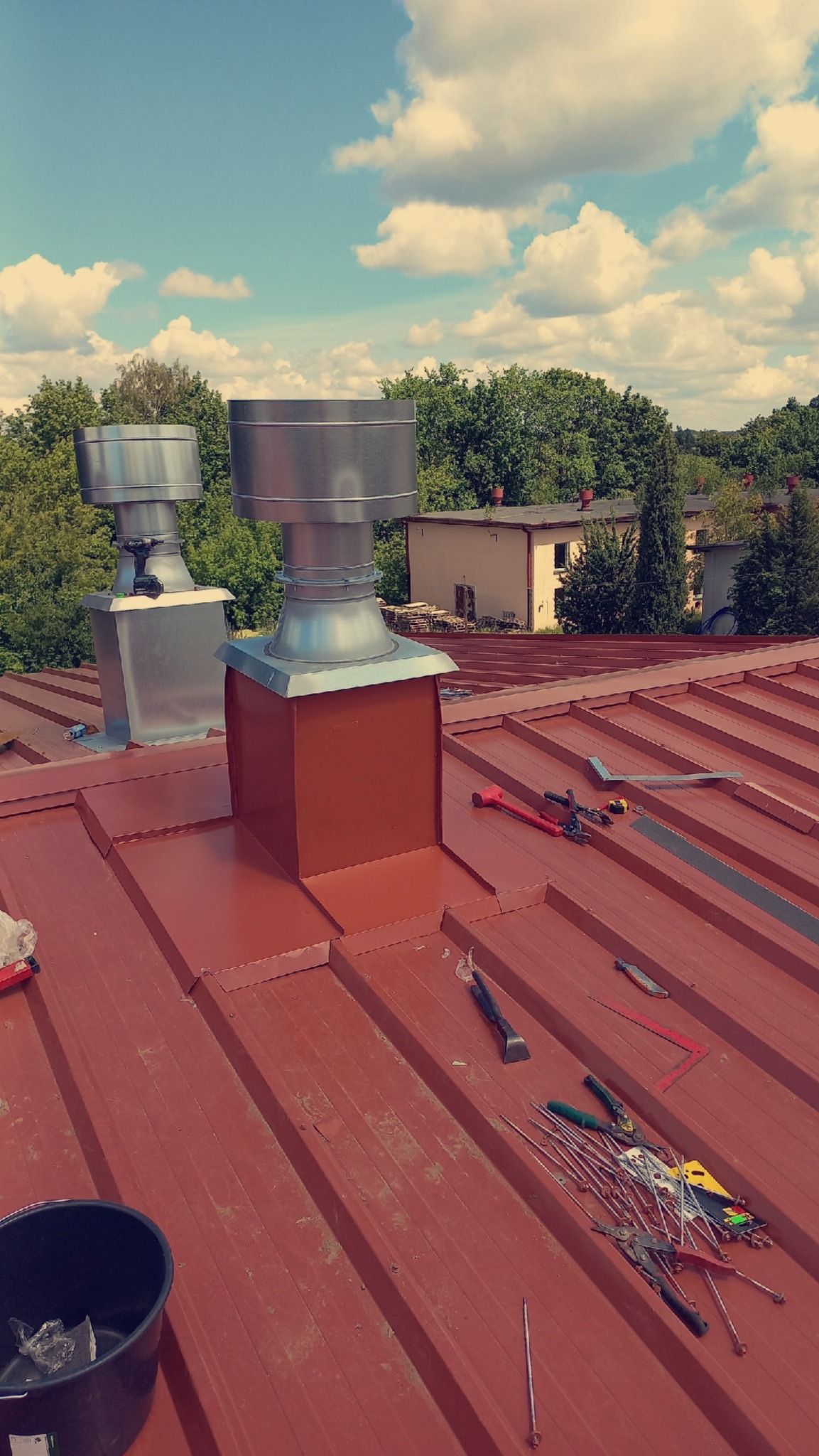 Czerwony dach z blachy trapezowej z dwoma kominami wentylacyjnymi i narzędziami dekarskimi. Widok z góry, słoneczny dzień, budynki i drzewa w tle.