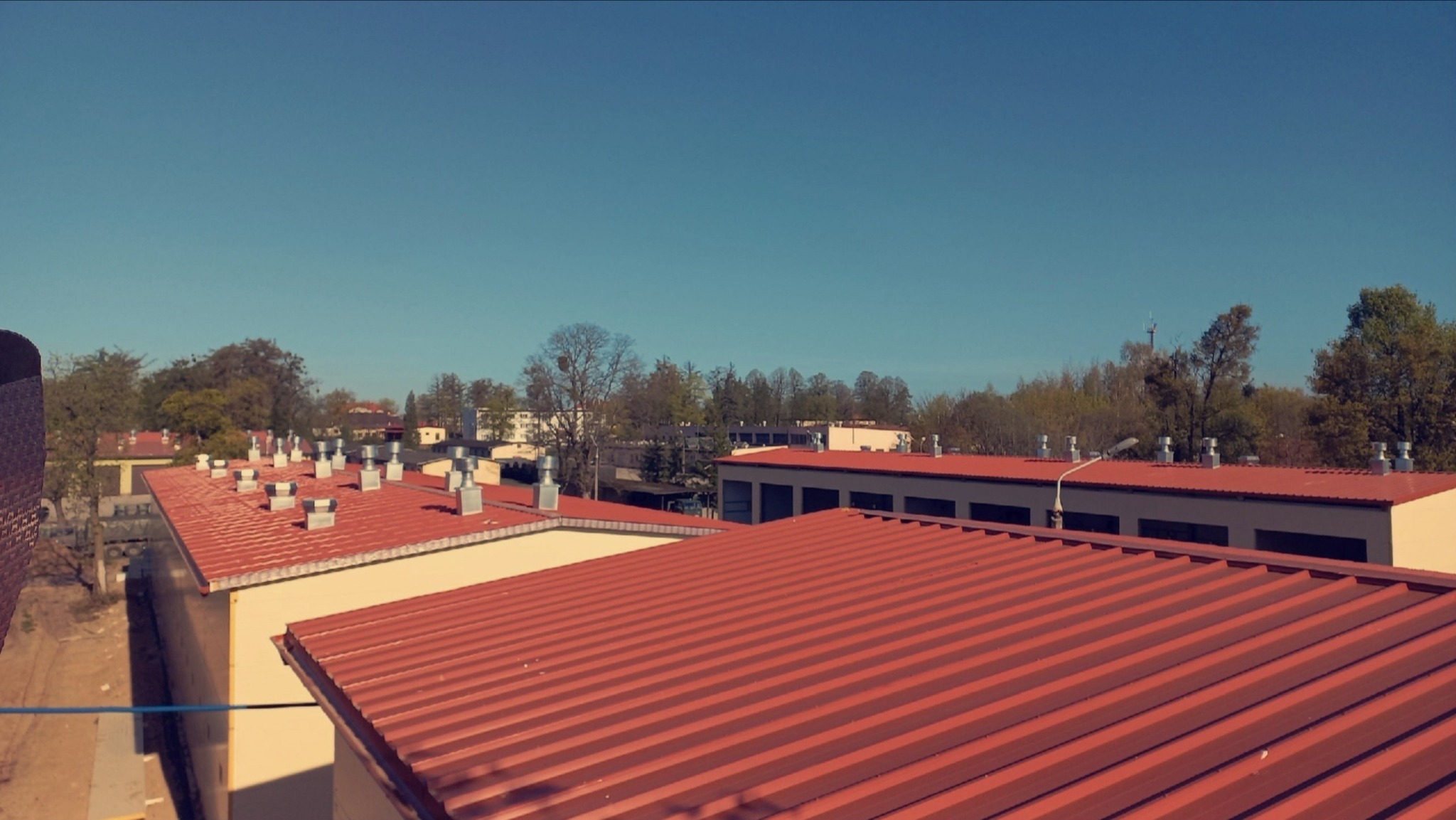 Czerwony dach z blachy trapezowej na budynku przemysłowym w słoneczny dzień. Widoczne kominy wentylacyjne i otoczenie z drzewami.