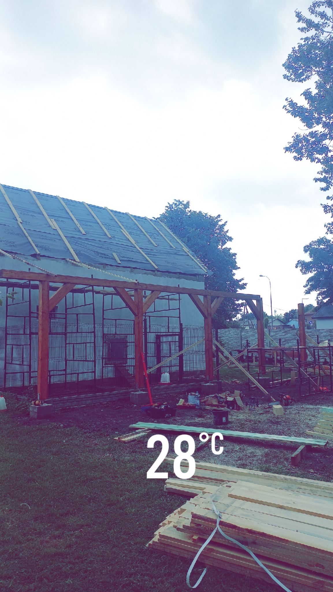 Wznoszenie konstrukcji dachu z drewnianą więźbą, pokrytej folią. W tle widoczny budynek i drzewa. Na pierwszym planie deski i trawa. Ujęcie z dołu.