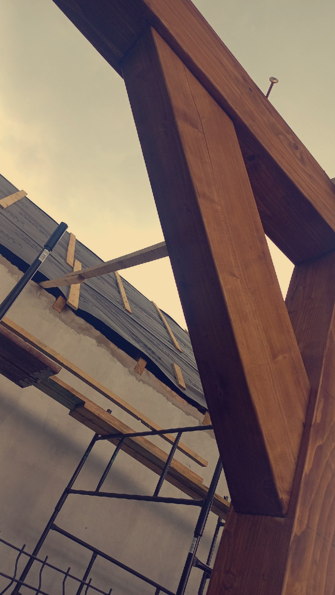 Wznoszenie więźby dachowej z drewnianych belek, widok z dołu na konstrukcję i fragment pokrycia dachowego na tle pochmurnego nieba.