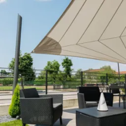 Nowoczesny taras z beżową, trójkątną osłoną przeciwsłoneczną, czarnymi meblami rattanowymi i zielonym trawnikiem, otoczony czarną balustradą.