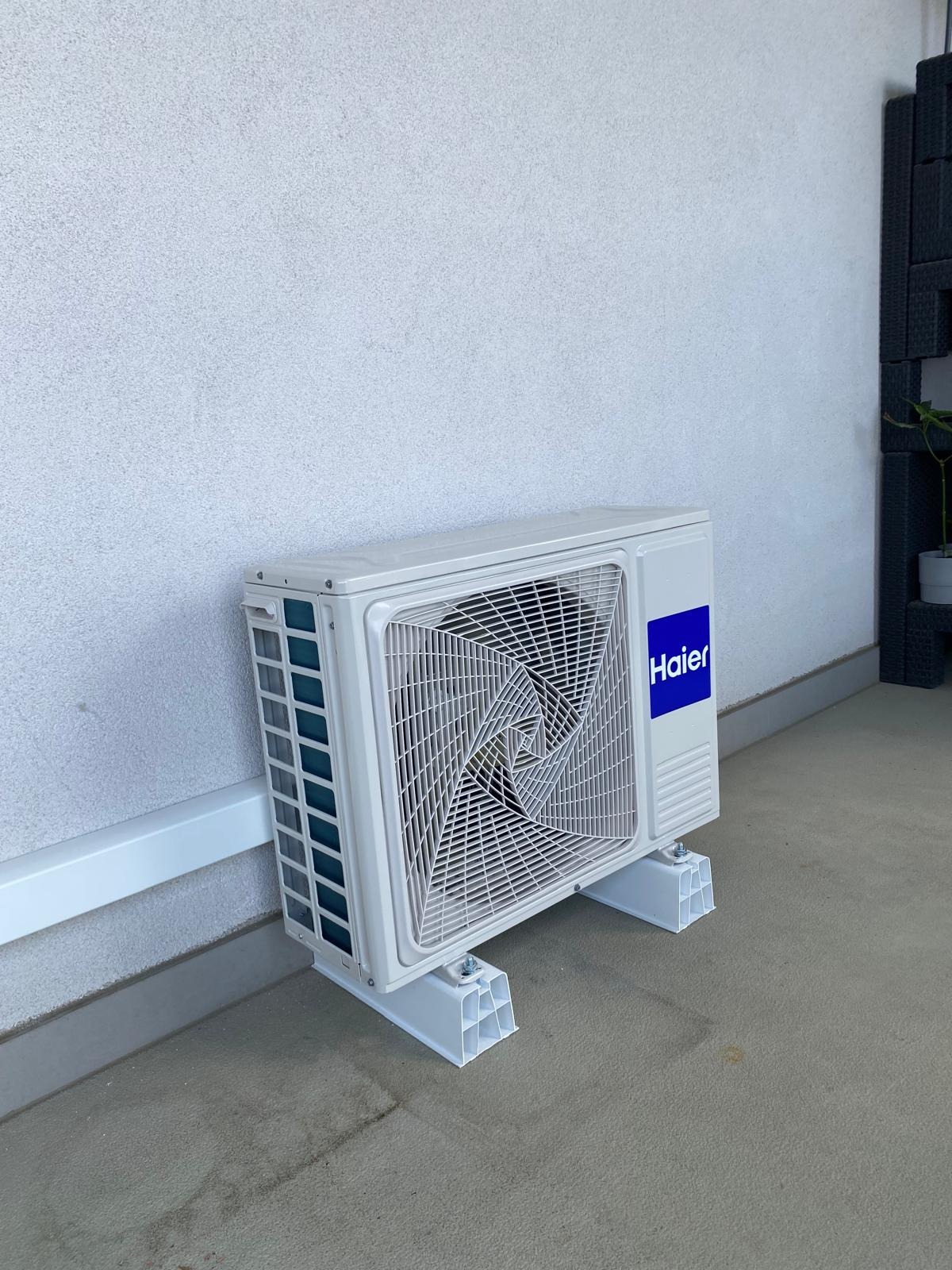 Biała jednostka zewnętrzna klimatyzacji Haier na plastikowych wspornikach, umieszczona przy jasnej, teksturowanej ścianie. Widoczne detale montażu i kratka wentylacyjna.