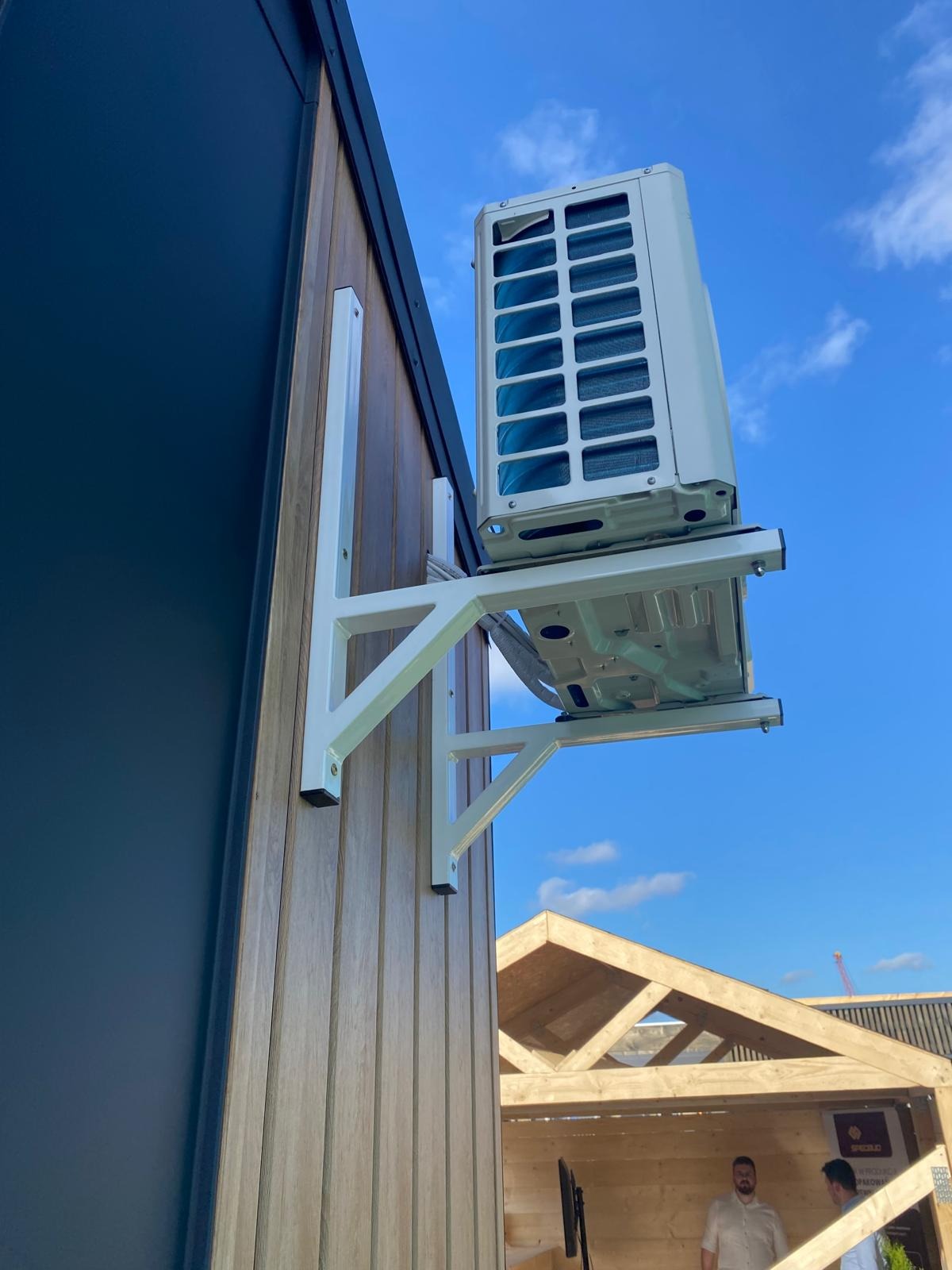 Biała jednostka zewnętrzna klimatyzacji zamontowana na drewnianej elewacji budynku, widok z dołu na błękitne niebo. Widoczne elementy konstrukcyjne budynku w tle.