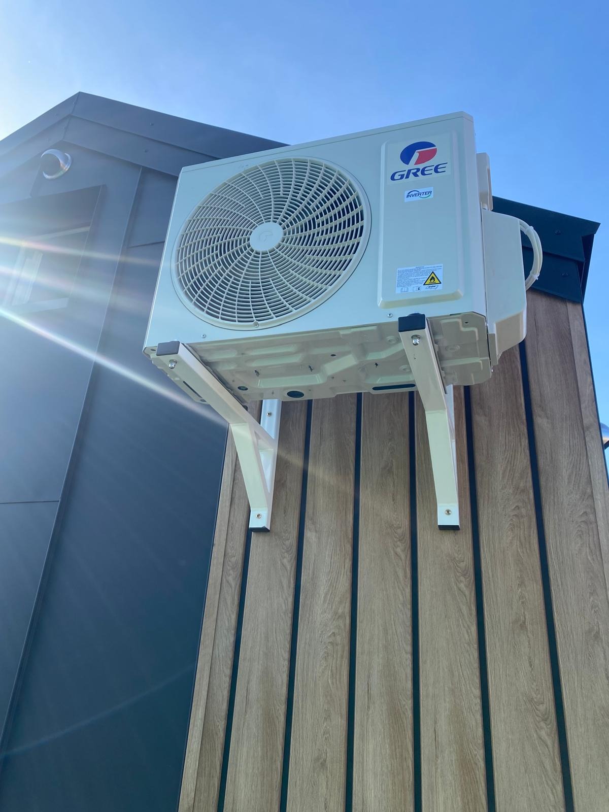 Zamontowany klimatyzator marki Gree na elewacji budynku z drewnopodobnymi panelami, widoczne białe wsporniki. Słońce odbija się od obudowy.