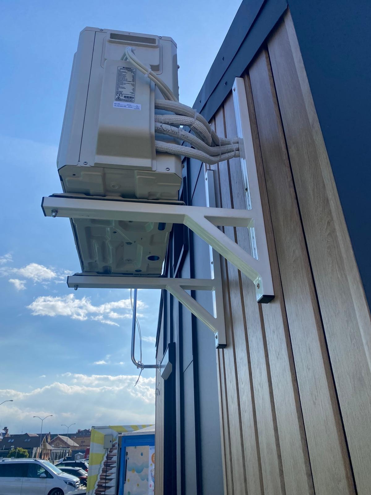 Zewnętrzna jednostka klimatyzacji zamontowana na elewacji budynku z drewnianą okładziną. Widoczne przewody połączeniowe i elementy mocujące.