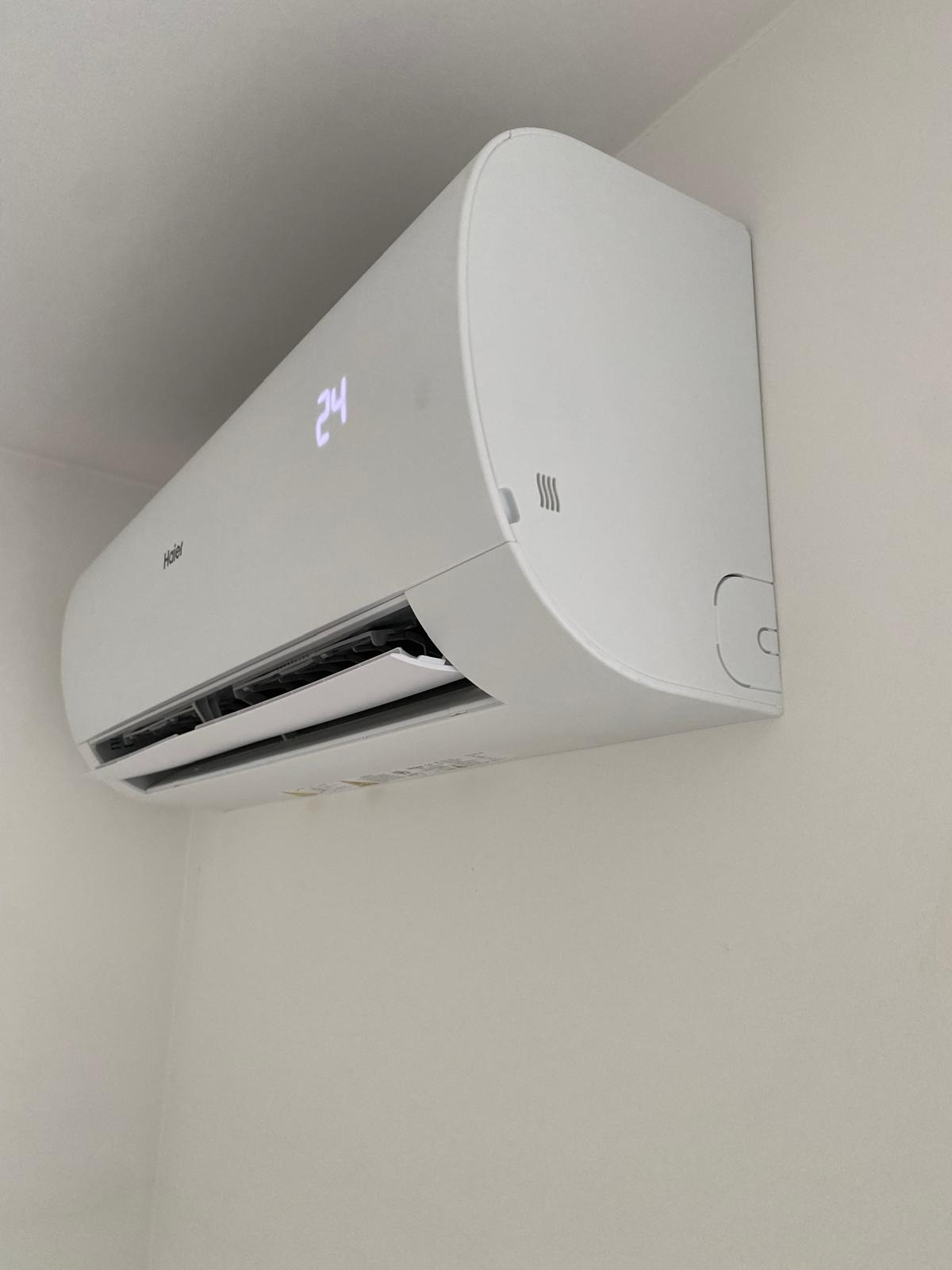 Biały klimatyzator Haier zamontowany na ścianie, wyświetlający temperaturę 24 stopnie Celsjusza. Widoczne żaluzje i logo producenta.