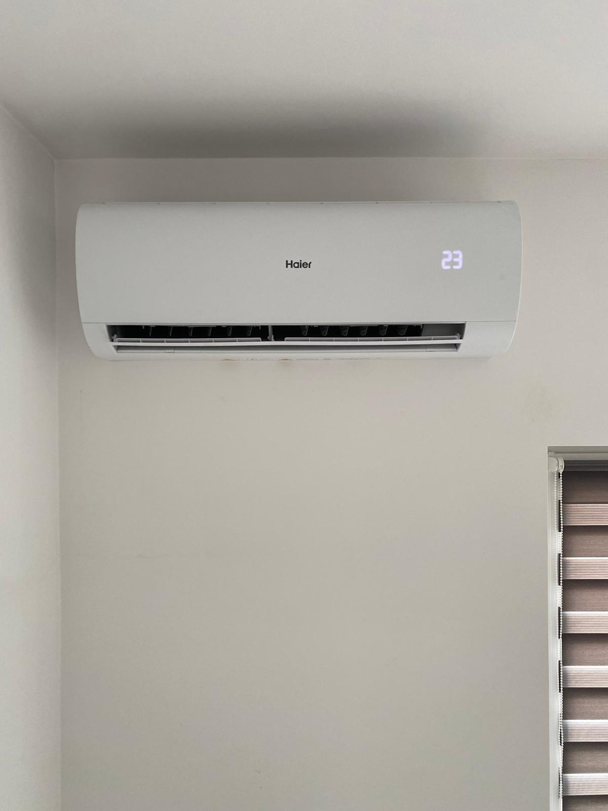 Biała klimatyzacja Haier zamontowana na ścianie. Na wyświetlaczu widoczna temperatura 23 stopnie. Obok fragment okna z roletami.