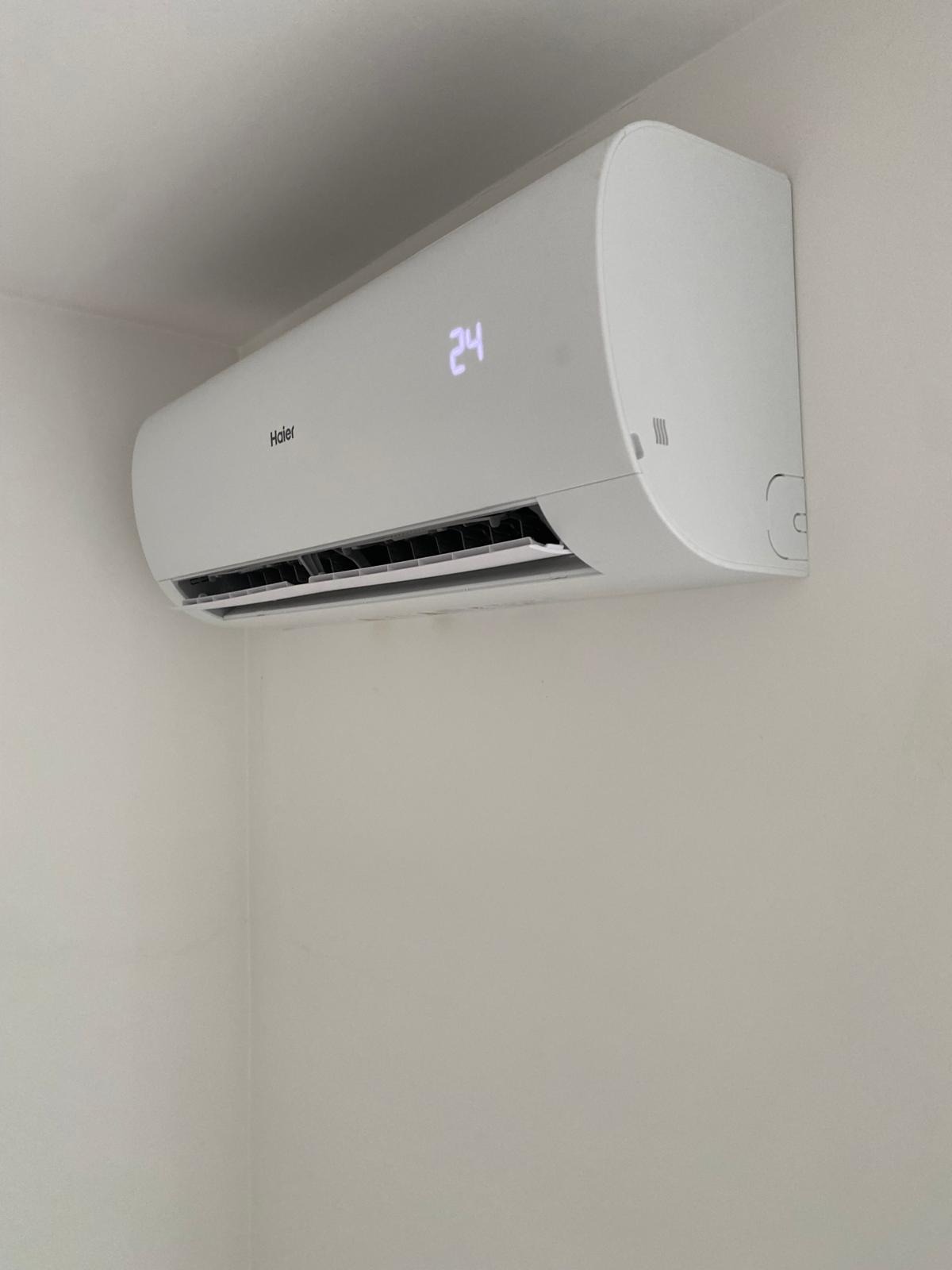 Biała, ścienna klimatyzacja Haier z wyświetlaczem ustawionym na 24 stopnie, zamontowana w rogu pomieszczenia o jasnych ścianach. Widoczne żaluzje wylotu powietrza.
