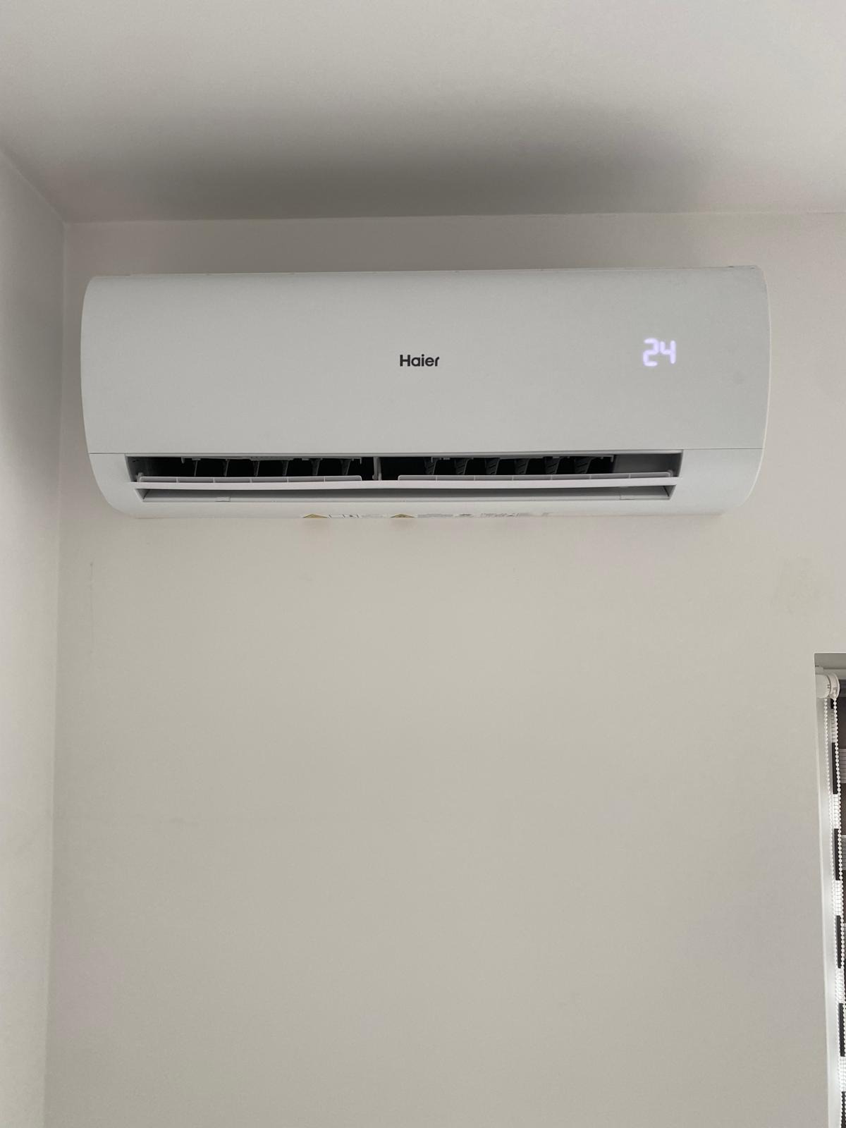 Biała klimatyzacja Haier na ścianie oświetlona naturalnym światłem, wyświetlająca temperaturę 24 stopnie Celsjusza. Widoczny fragment rolety okiennej.