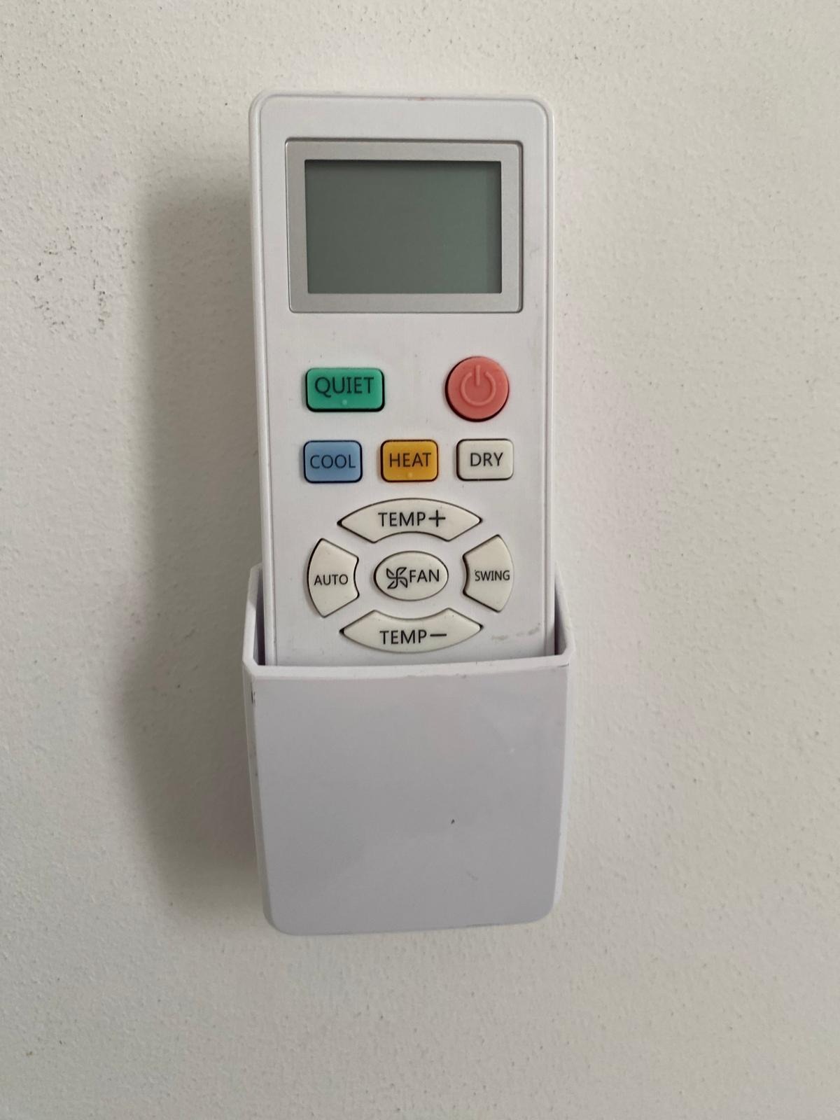 Biały pilot do klimatyzacji z kolorowymi przyciskami (Quiet, Cool, Heat, Dry, Temp) zamontowany na ścianie w uchwycie.