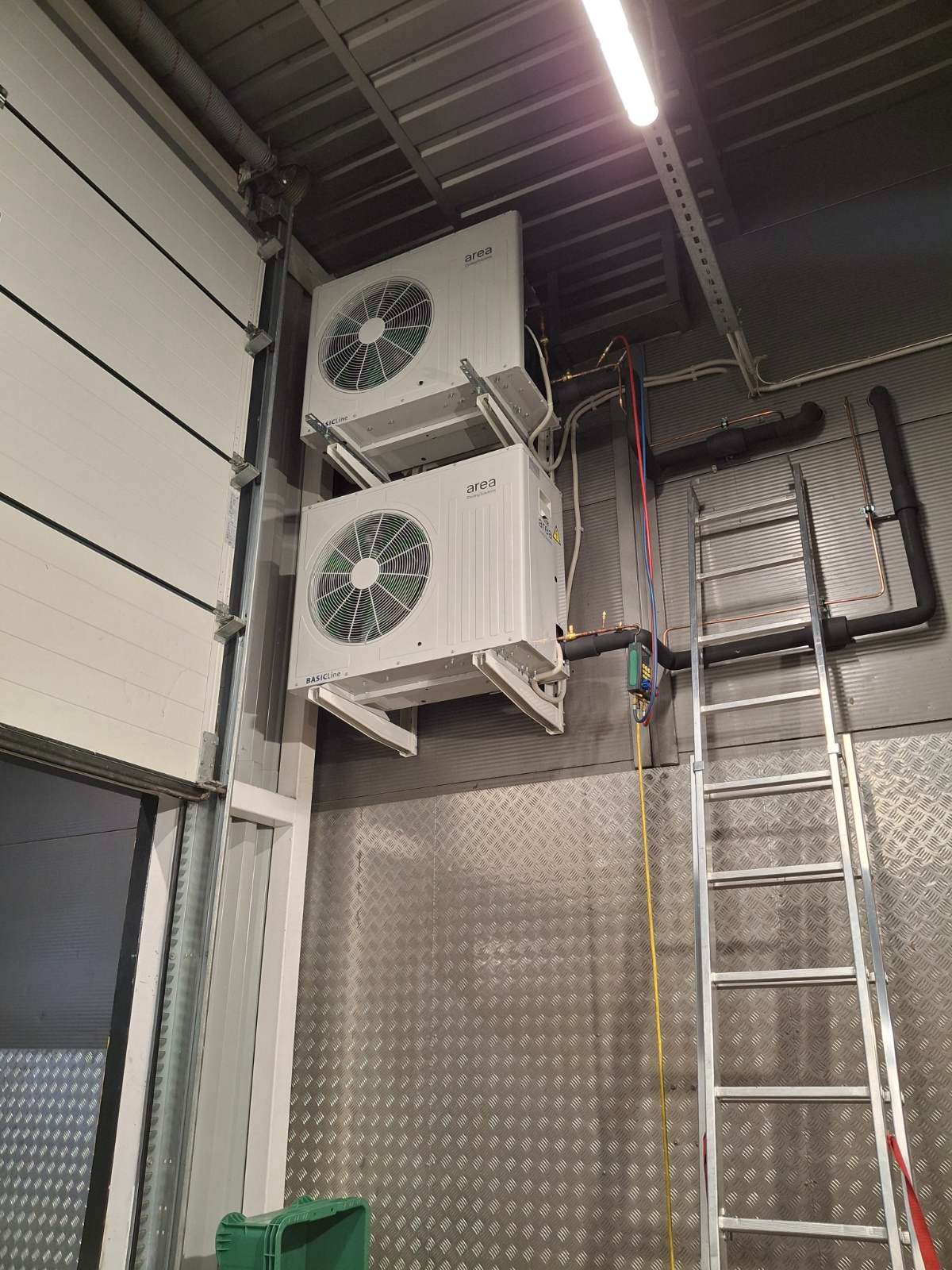 Dwa agregaty klimatyzacyjne AREA, zamontowane jeden nad drugim na ścianie z blachy trapezowej w hali, obok drabina aluminiowa i instalacja rurowa.
