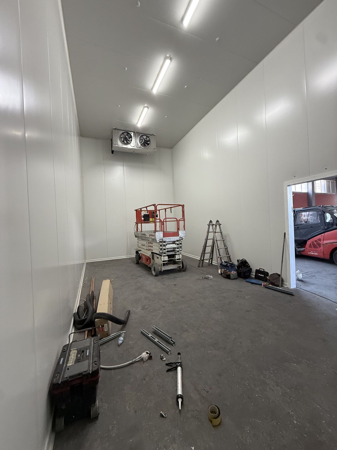 Wnętrze chłodni z zamontowaną klimatyzacją przemysłową, widoczny podnośnik i narzędzia instalacyjne. Białe panele ścienne i sufitowe, betonowa posadzka.