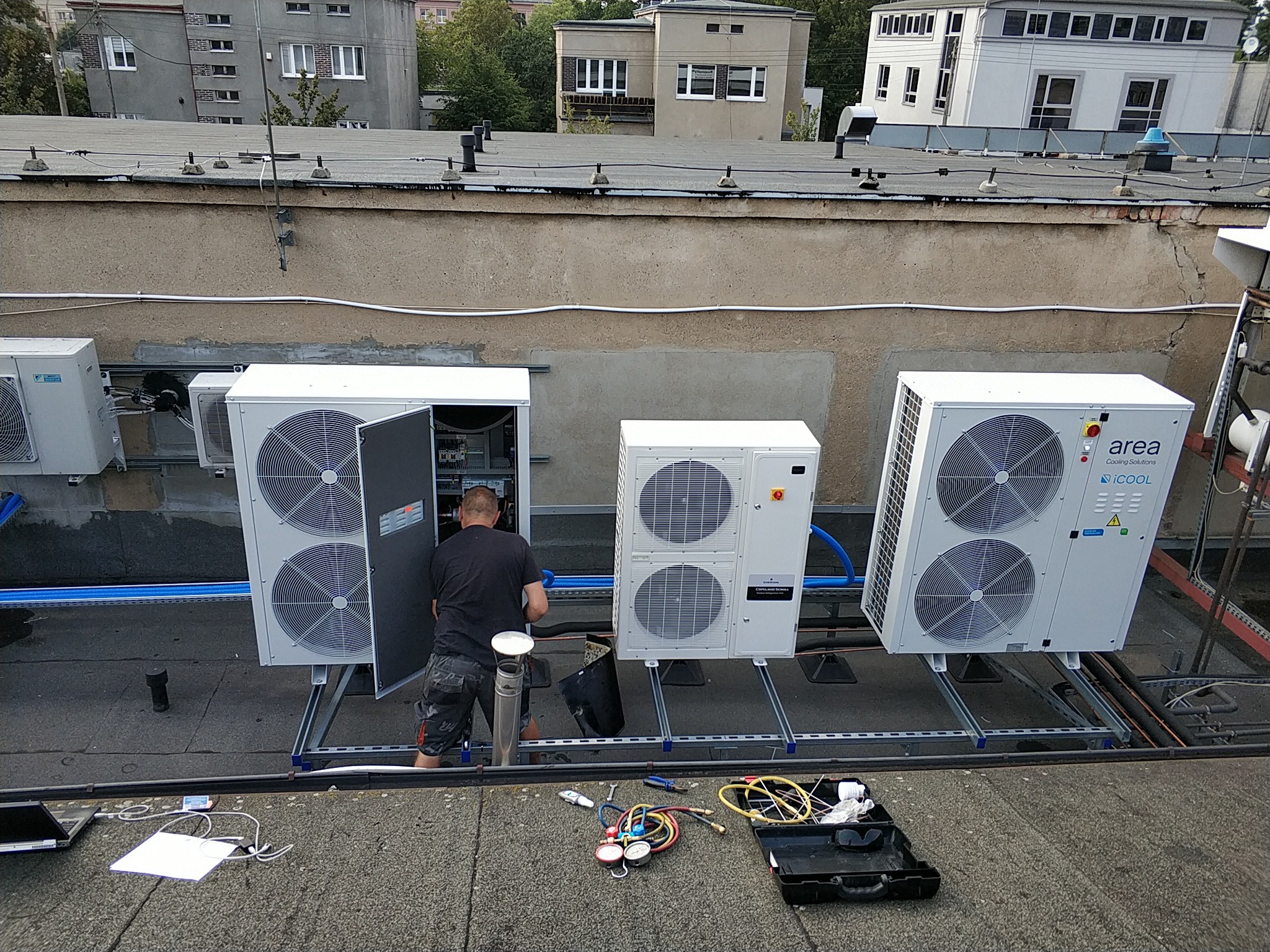 Serwisant w trakcie naprawy klimatyzacji na dachu budynku. Widoczne jednostki zewnętrzne klimatyzatorów, narzędzia pracy i elementy instalacji chłodniczej.