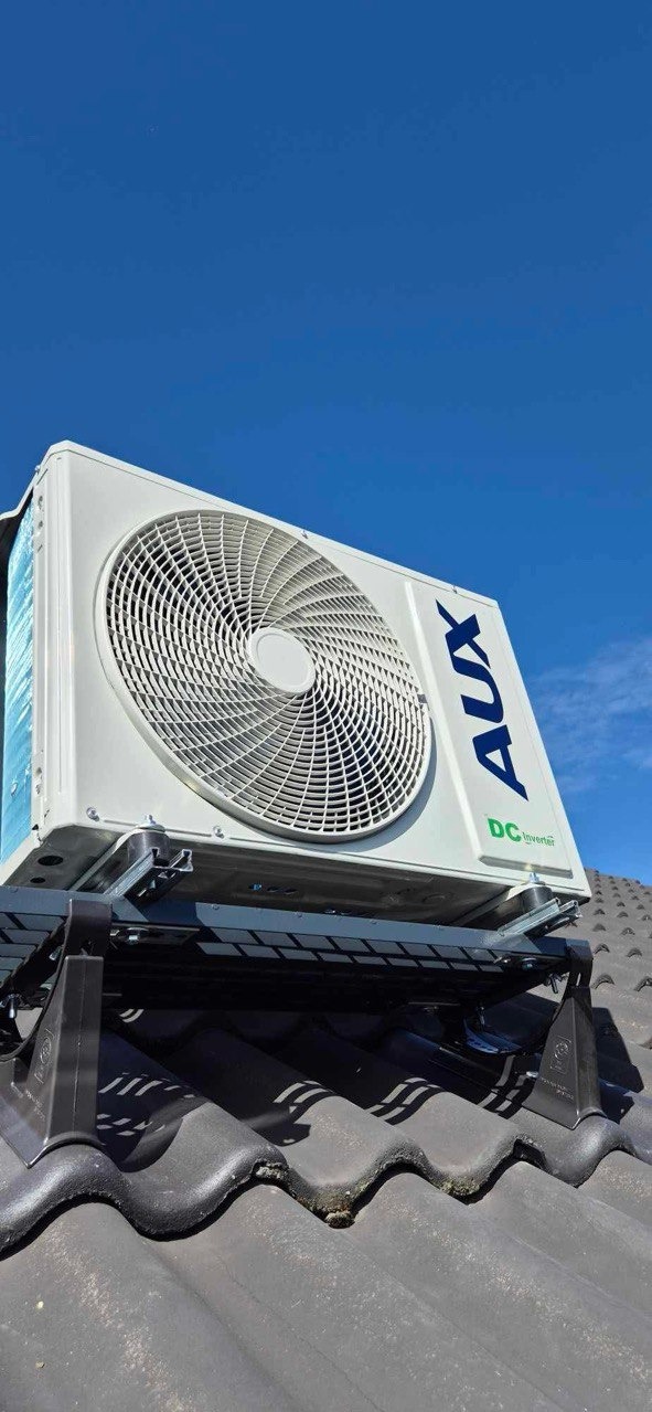 Biała jednostka zewnętrzna klimatyzacji AUX, zamontowana na dachu z czarnej dachówki, na specjalnej czarnej konstrukcji wsporczej. Błękitne niebo w tle.