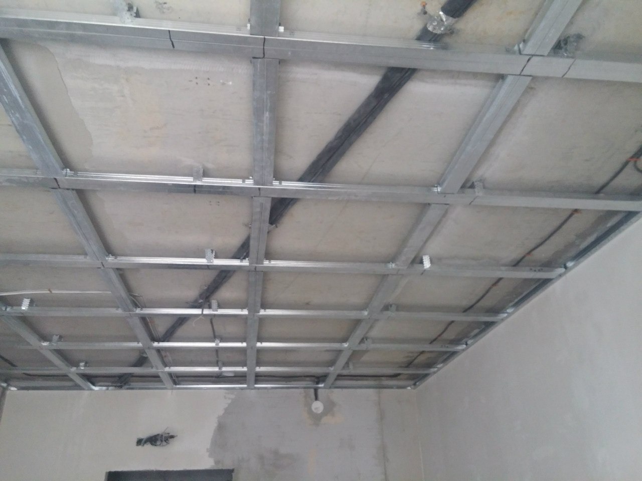 Stelaż sufitu podwieszanego z widocznymi przewodami instalacji, przygotowany do montażu płyt. Widok z dołu, surowy stan wykończenia wnętrza.