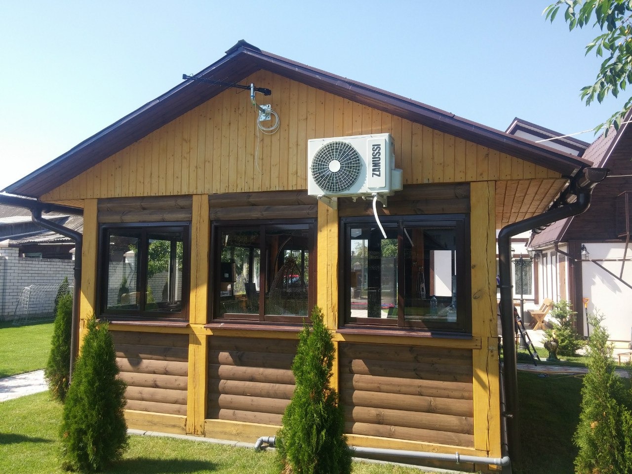 Zewnętrzny klimatyzator Zanussi zamontowany na drewnianej altanie z brązowymi oknami, widok z ogrodu w słoneczny dzień.