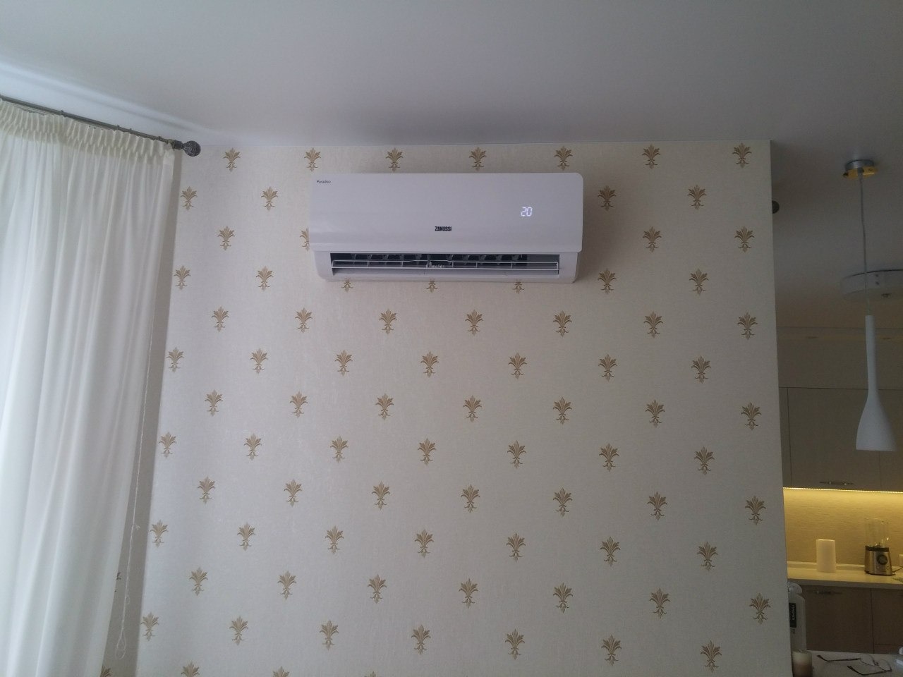 Biały klimatyzator Zanussi zamontowany na ścianie z jasną tapetą w delikatne, złote wzory. Widoczny fragment zasłony i lampa wisząca.