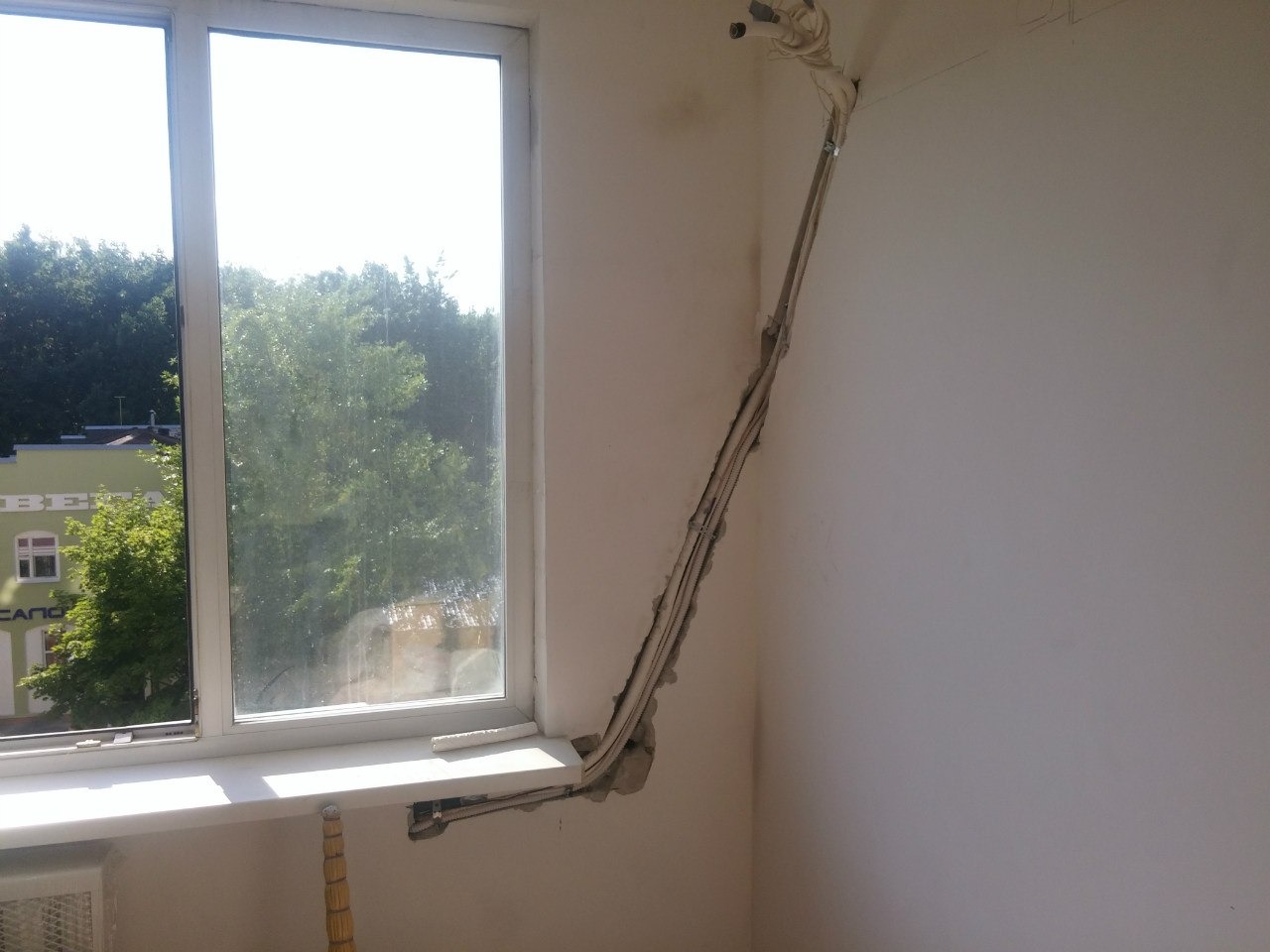 Montaż instalacji klimatyzacji w pomieszczeniu: kable i rury prowadzone po ścianie, widoczne elementy montażowe. Przez okno widać zieleń drzew.