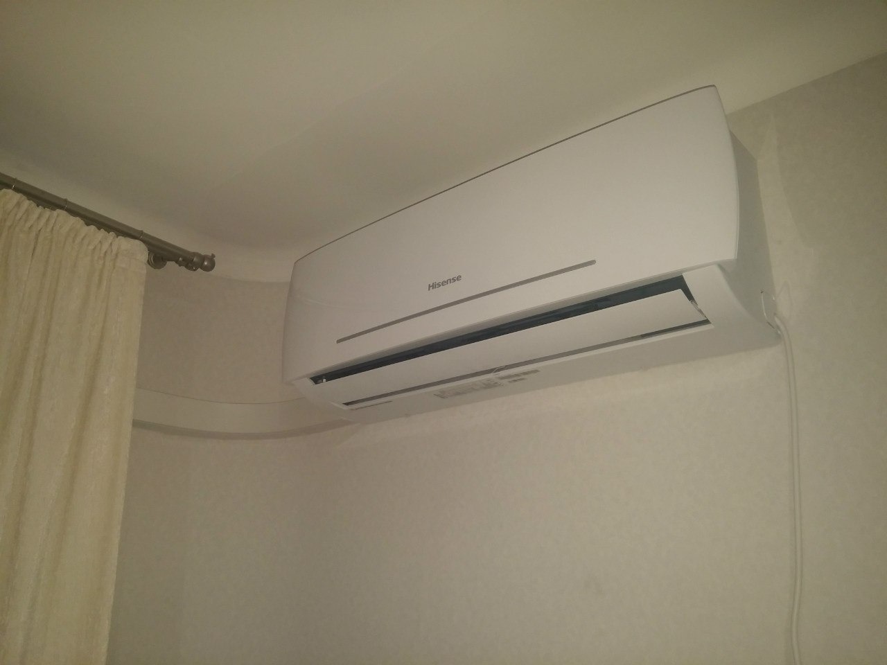 Biała klimatyzacja Hisense zamontowana na ścianie w pokoju, widoczna krótka firanka i kabel zasilający. Ujęcie z dołu, lekko z prawej strony.